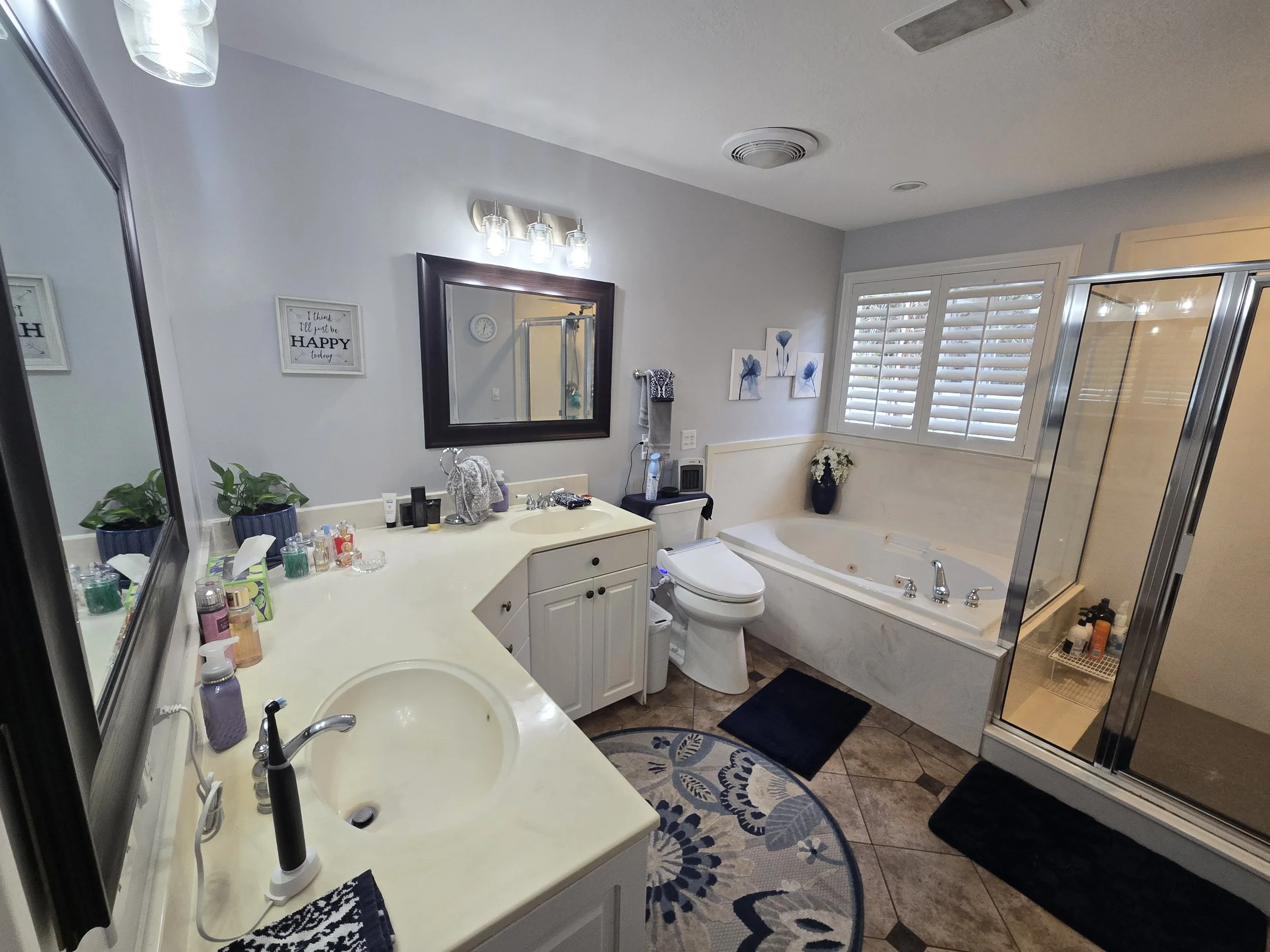 A bathroom with white vanity, two sinks, various toiletries, a large mirror, artwork of blue flowers, a window with white blinds, a bathtub with a black vase holding white flowers, a shower enclosure, and bathroom accessories including towels, a cloc