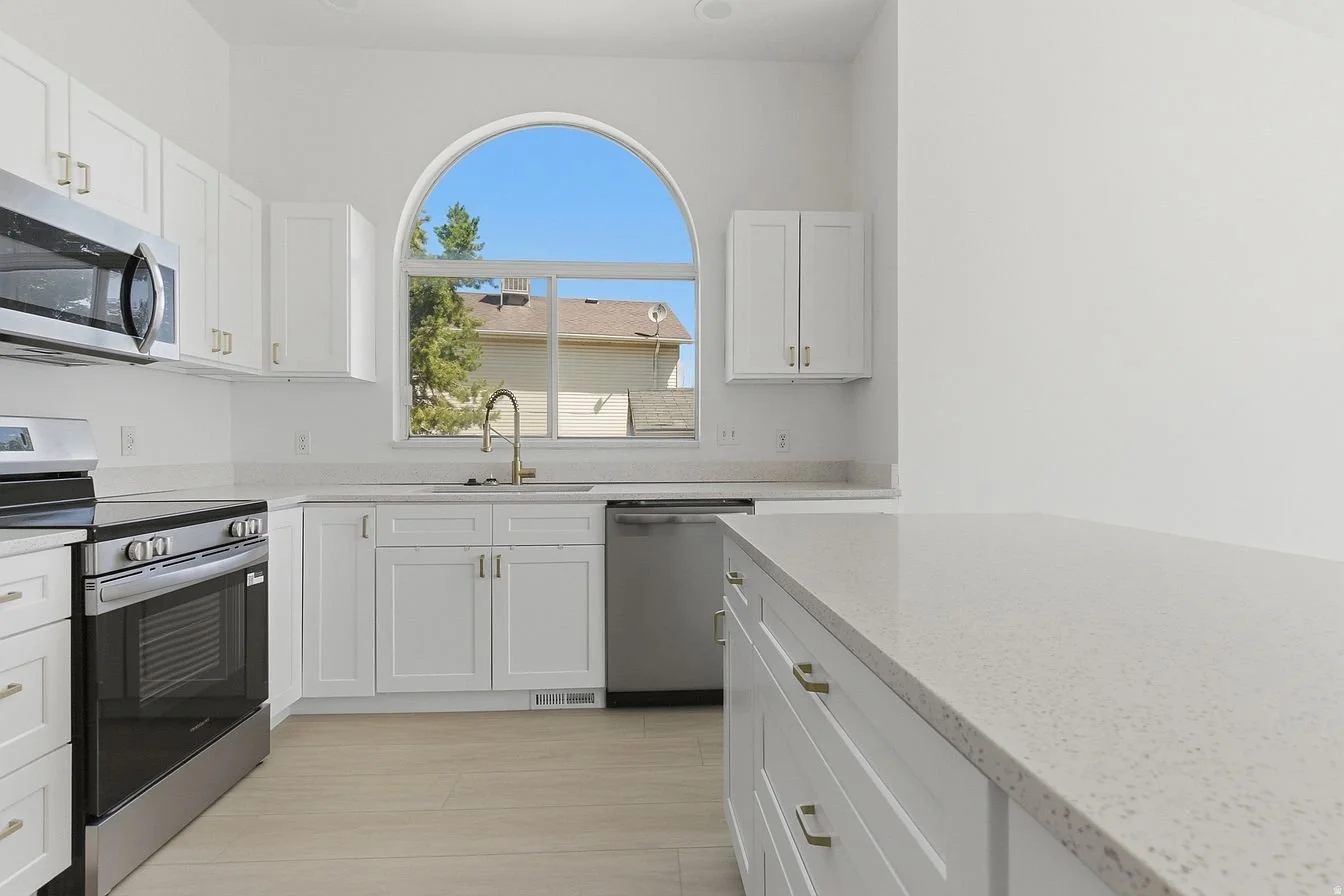 Empty kitchen with white cabinets, a large arched window, and stainless steel appliances, including a stove, microwave, and dishwasher.