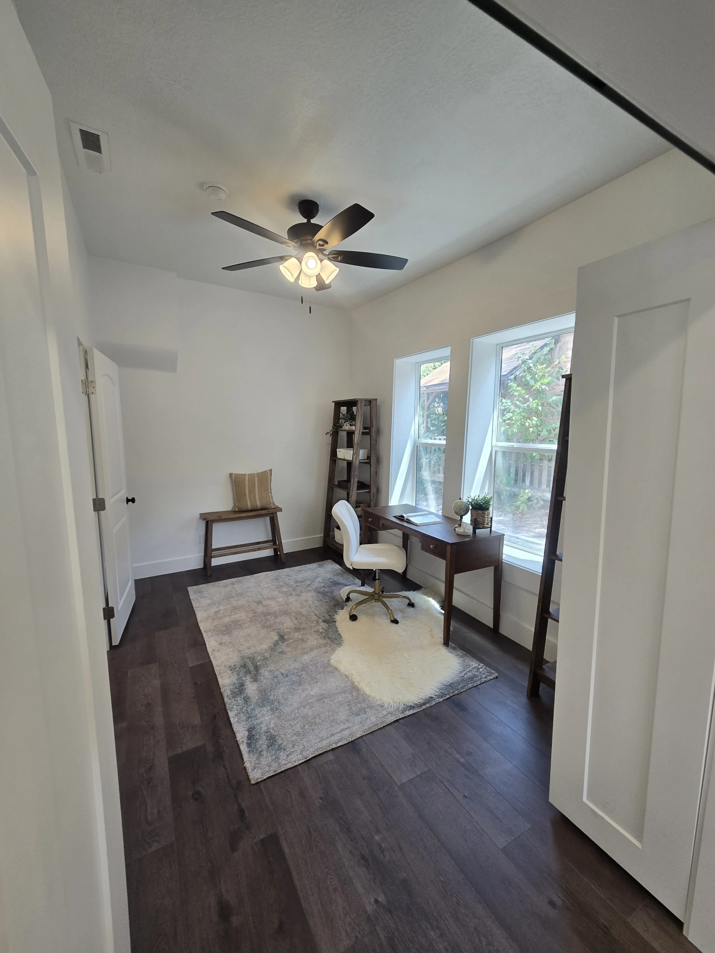 A neat and modern home office with a dark wood floor, a white rug, a small wooden desk with a globe and plants, a white desk chair, a wooden ladder shelf with decor, a wall with large windows, a ceiling fan with lights, and a chair with a striped cus