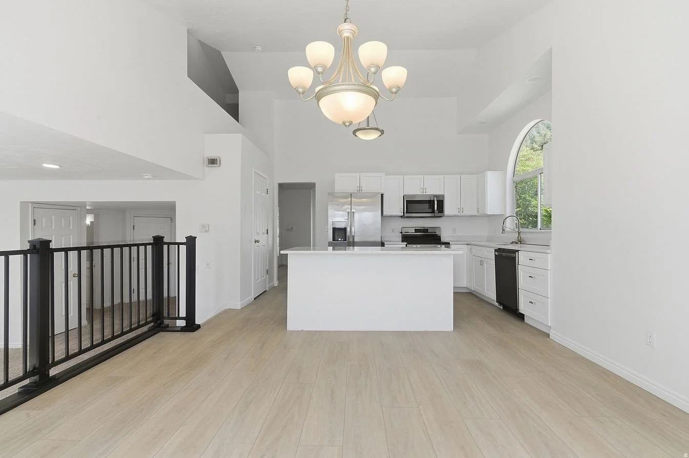 Empty modern white kitchen with island, stainless steel appliances, large arched window, and a chandelier, with light wood flooring.