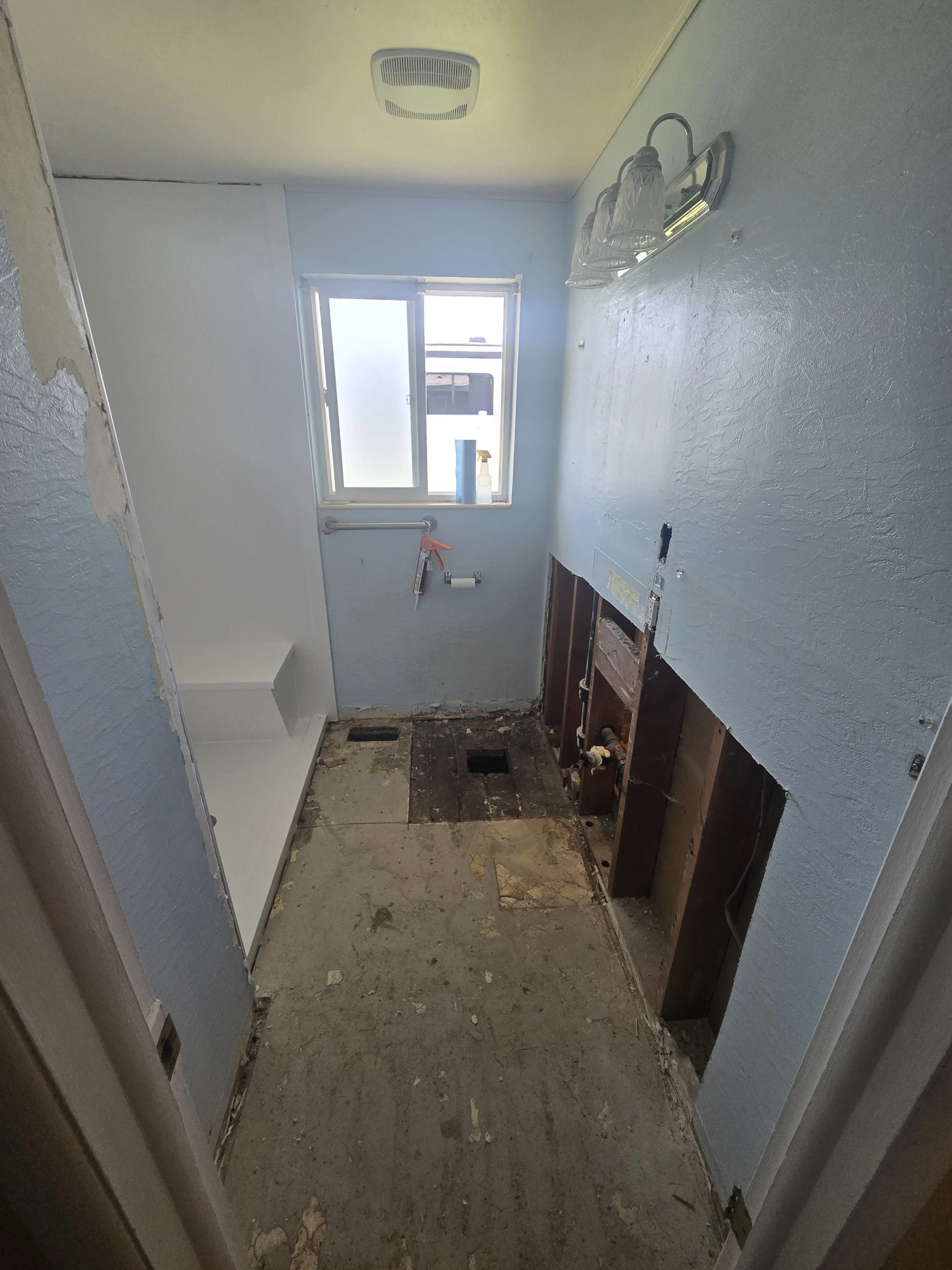 A small bathroom under renovation with exposed floor and wall framing, a partially removed vanity, a window, and a ceiling vent.