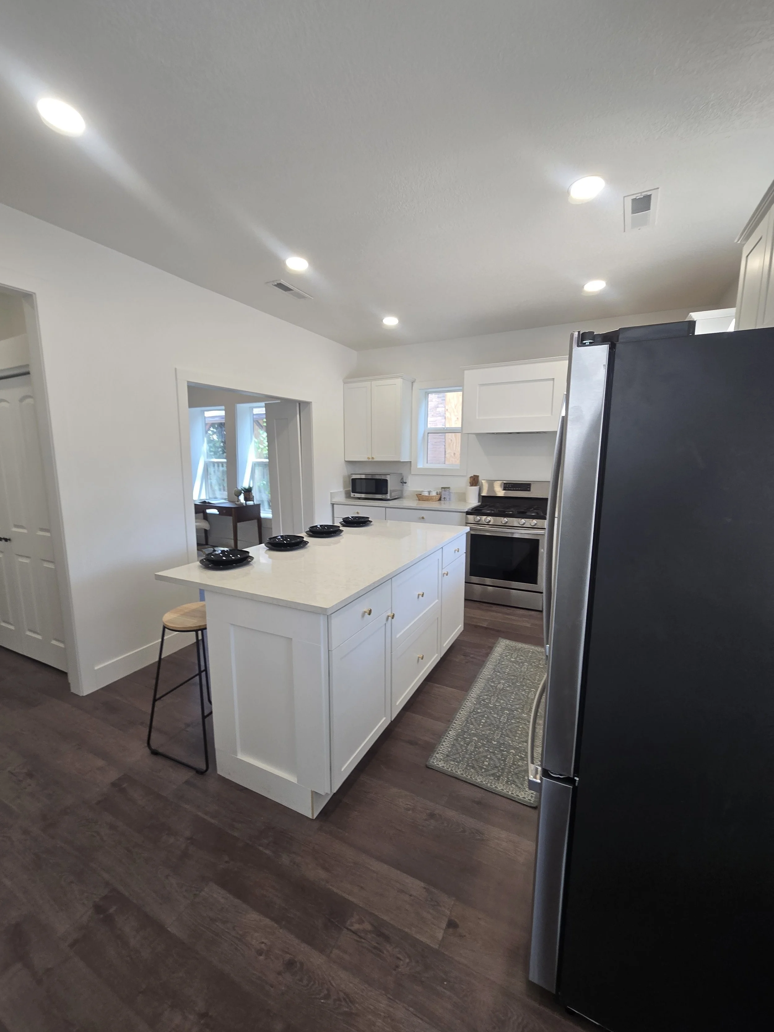 A modern kitchen with dark hardwood flooring, white cabinets, a white island with gold handles, and a stainless steel refrigerator. There is a window above the sink allowing natural light, and a small round stool next to the island.