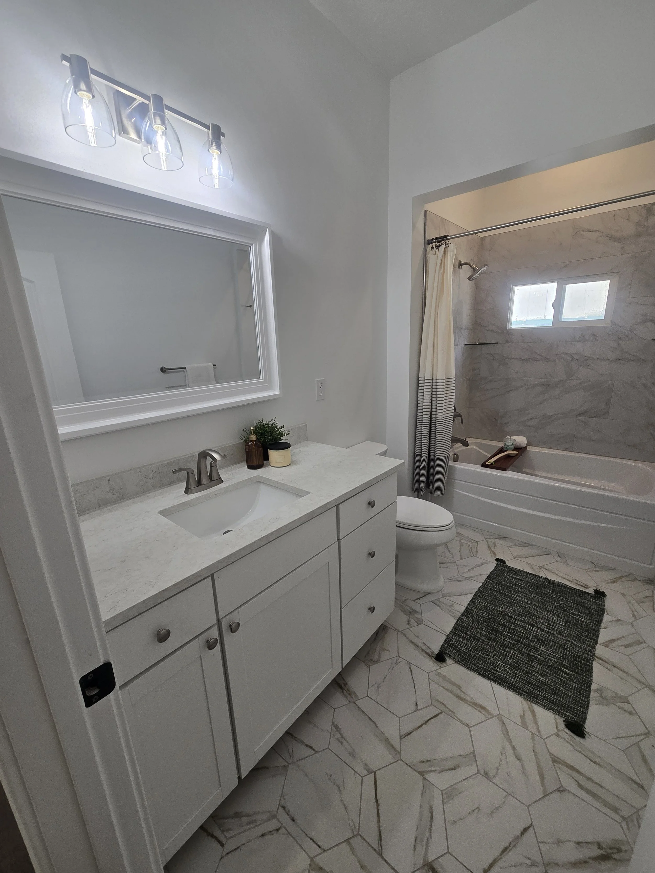 A bathroom with a white vanity, marble countertop, large mirror, three light fixtures, a toilet, shower with a small window, towel bar, black rug with tassels, and marble tile flooring.