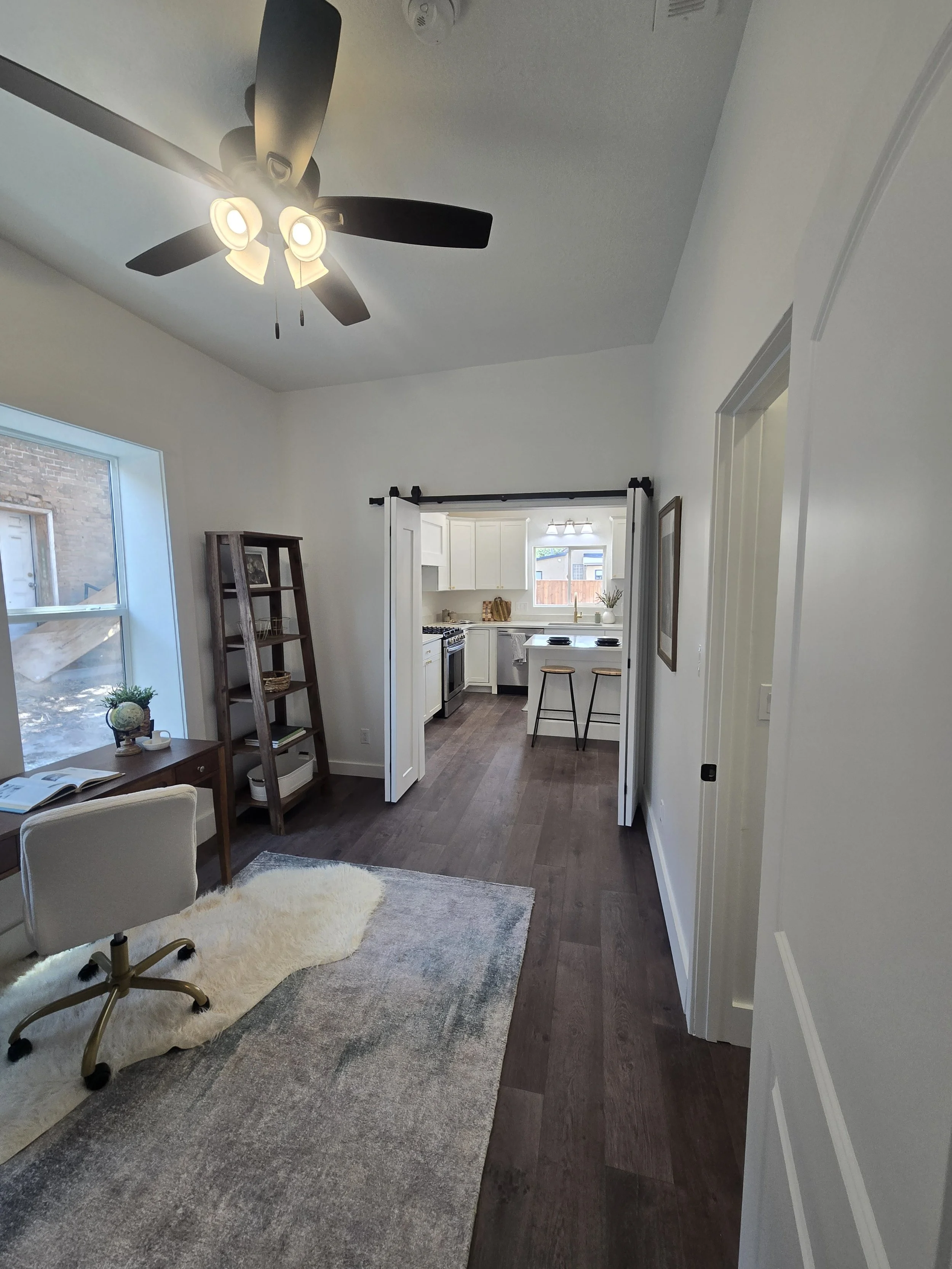 Interior view of a living room leading into a kitchen with white cabinets, a window, and a black countertop with stools. A ceiling fan with lights is on the ceiling. There is a desk with an open book, a chair, a small bookshelf, and a floor rug in th