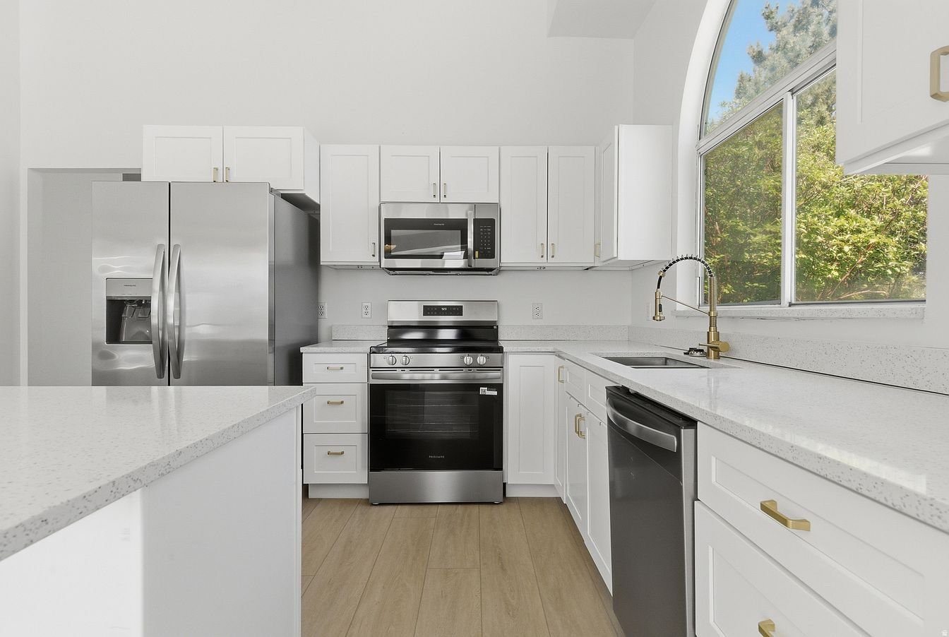 Modern kitchen with white cabinets, stainless steel appliances, including refrigerator, microwave, oven, and dishwasher. A large window with a view of trees and blue sky.
