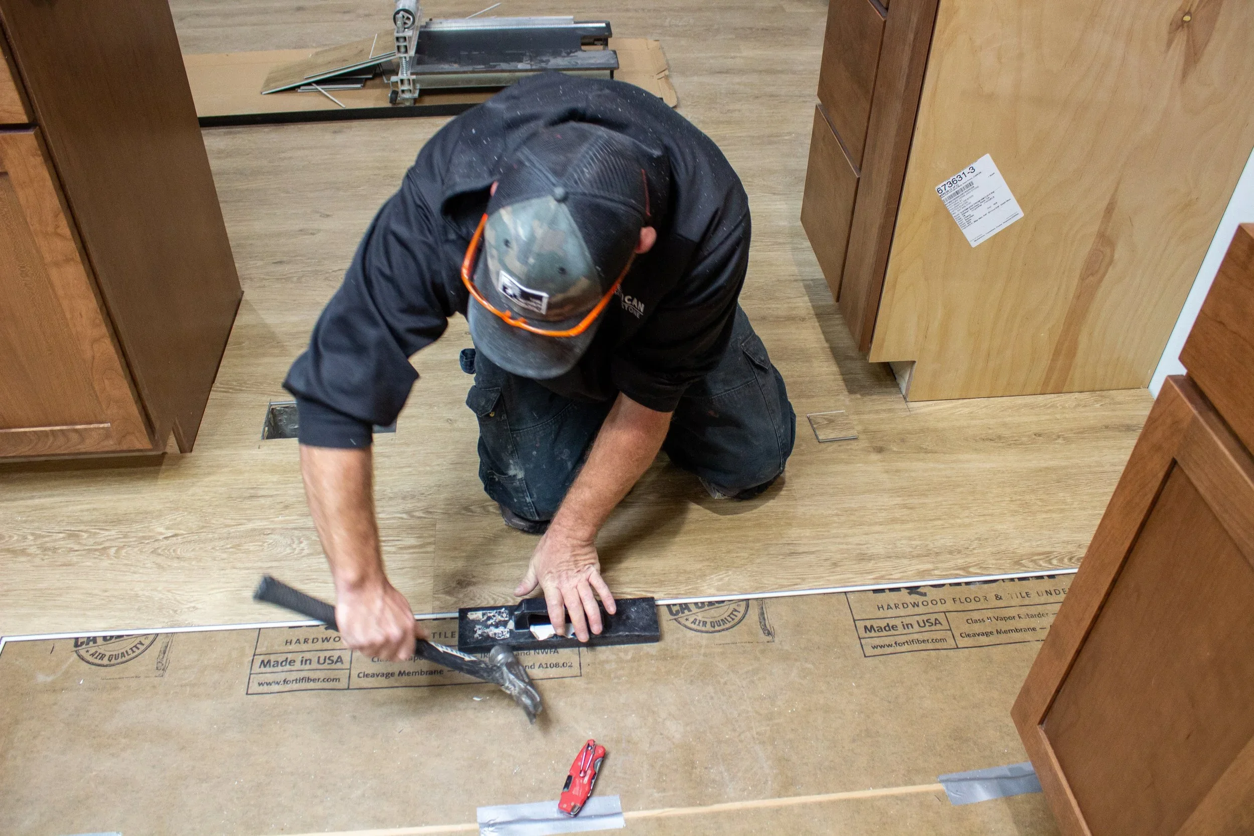A man in a black shirt, baseball cap, and orange safety glasses kneels on the floor, installing or working on wooden flooring in a room with wooden cabinets.