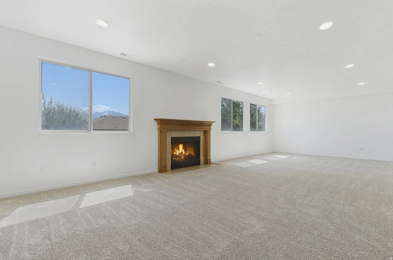 Empty living room with white walls, beige carpet, a fireplace with a wooden mantel, three windows, and recessed ceiling lights.