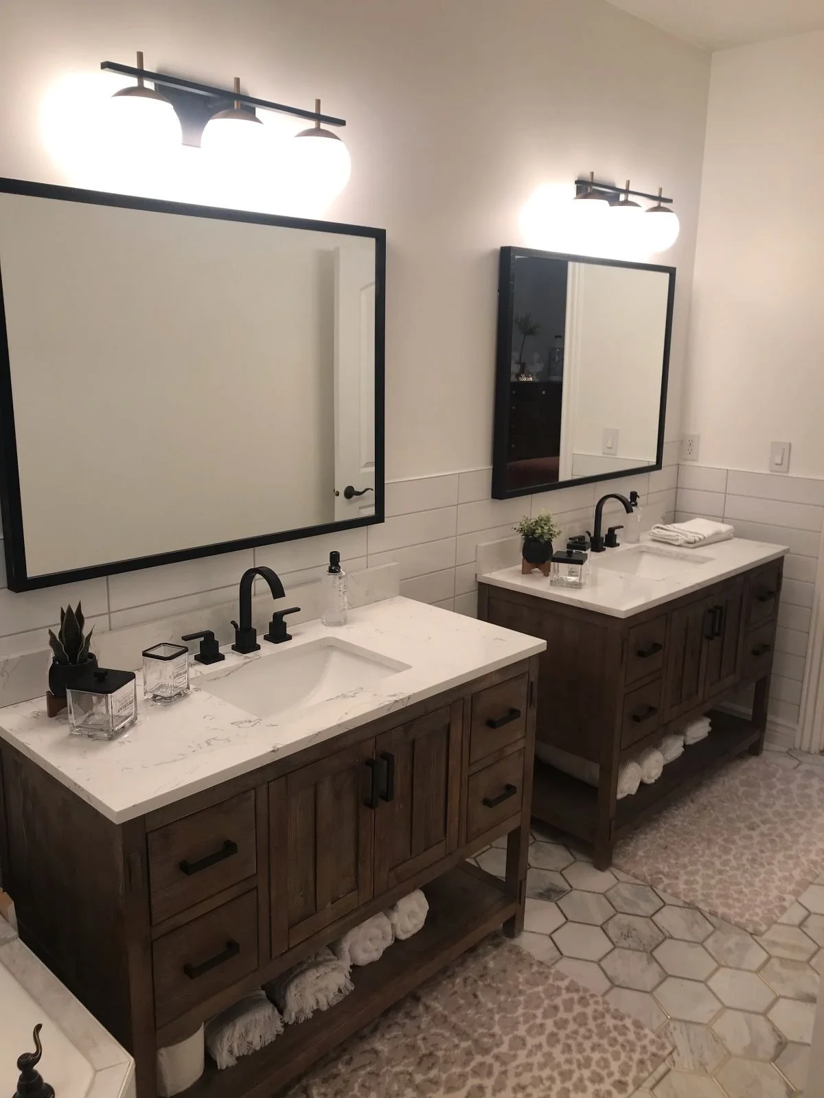 Double bathroom vanity with white marble countertops, wooden cabinets, black faucets, and two large rectangular mirrors with black frames. Above each mirror, there are modern light fixtures. The vanity top has small plants, soap dispensers, and towel