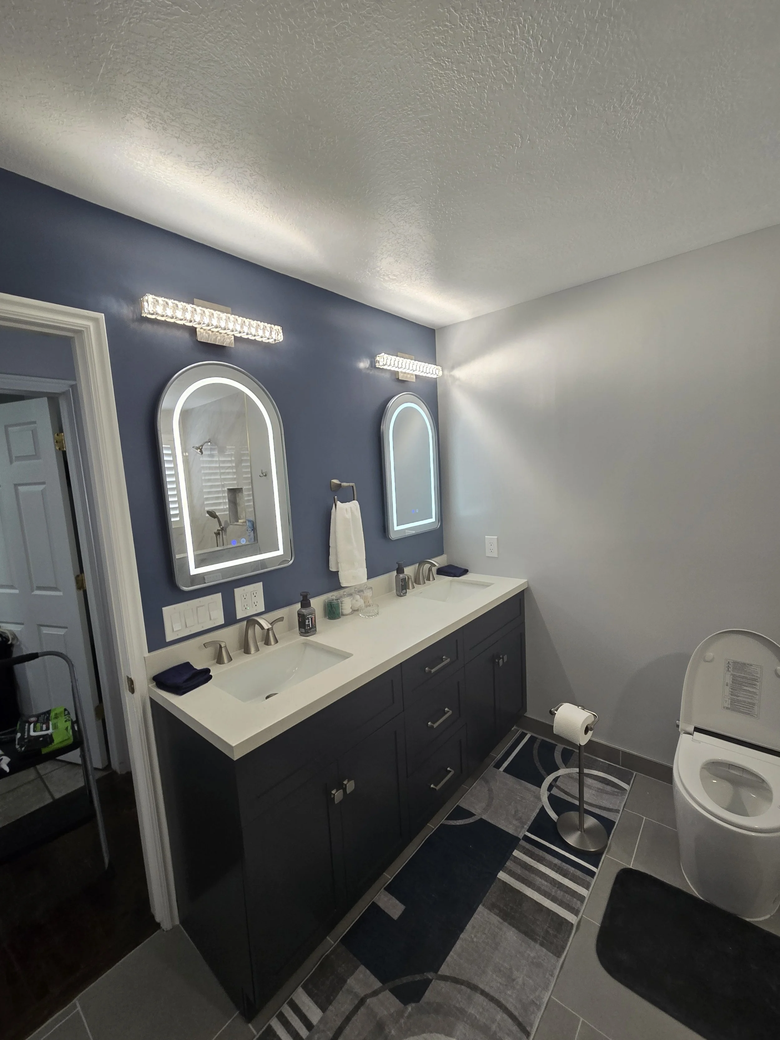 A bathroom with a dark blue vanity, two illuminated mirrors, a double sink, and a toilet. The bathroom has dark and light gray tiled floors, a patterned rug, and a small step stool.