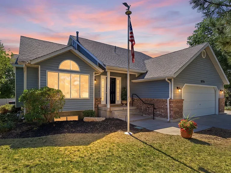 Front view of a blue house with a porch, large window, and attached garage, during sunset with a colorful sky, American flag on a pole, and a potted plant on the lawn.