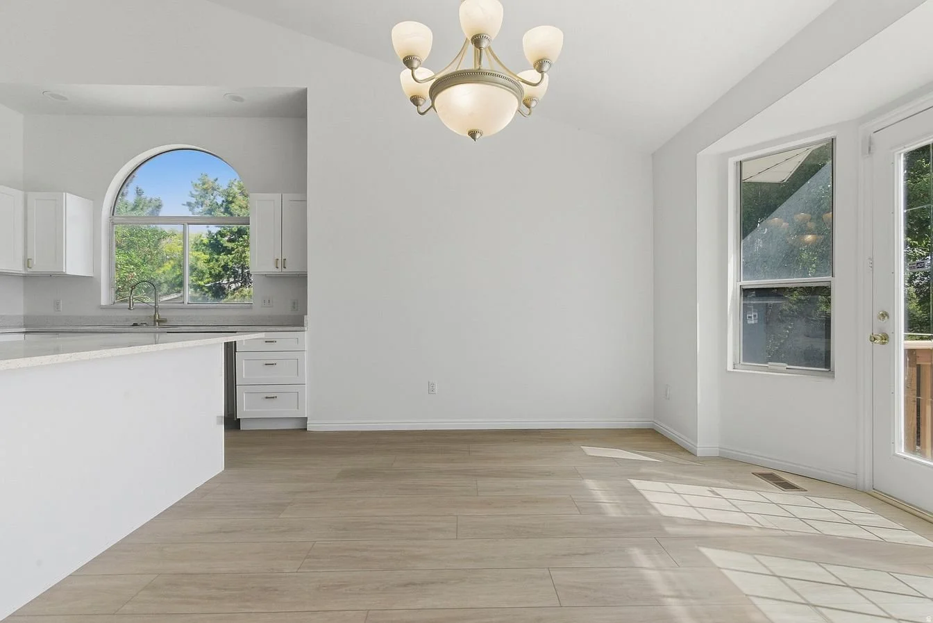 Empty dining area adjacent to the kitchen with a chandelier and large windows showing greenery outside.