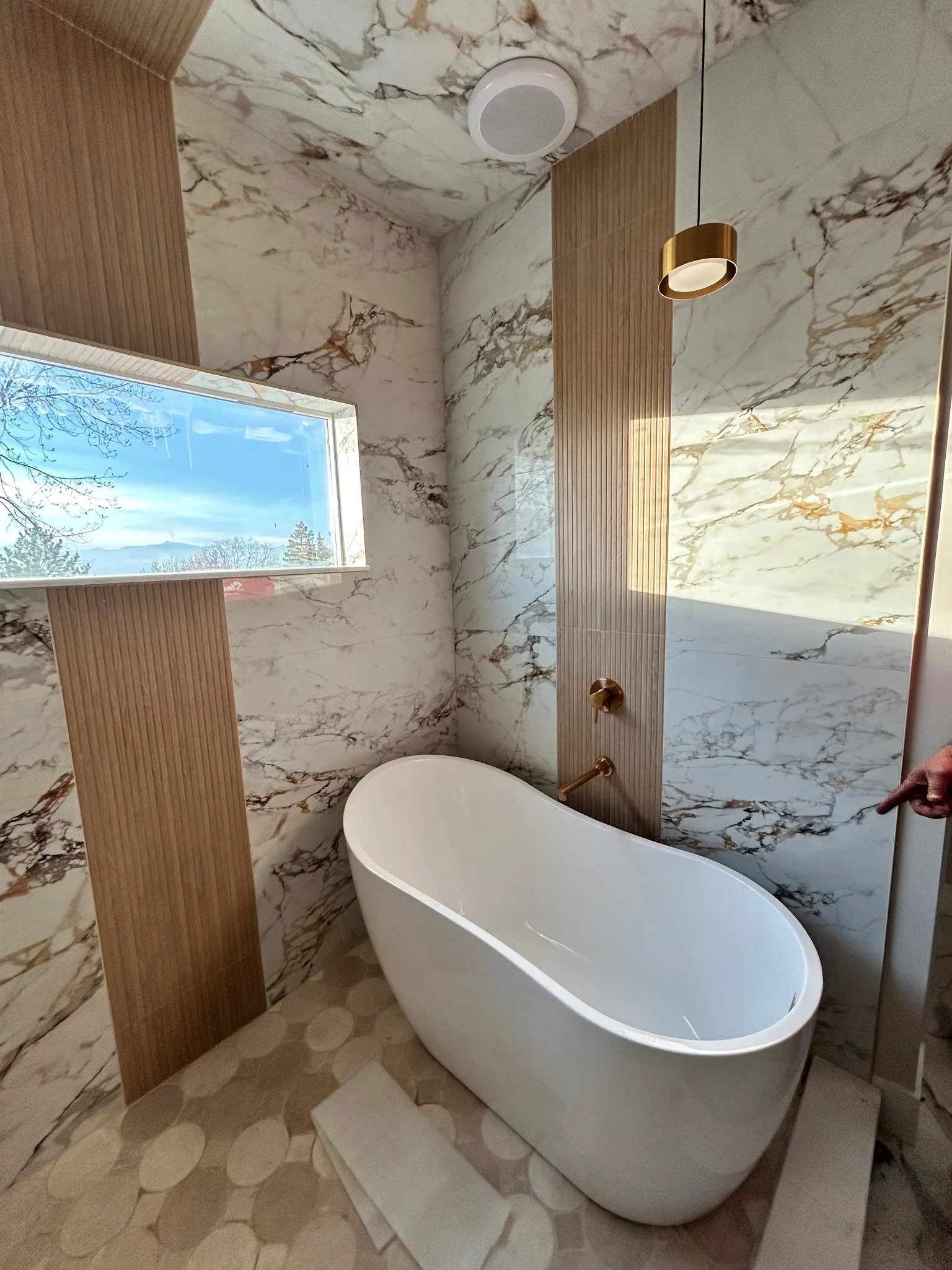 A modern bathroom featuring a freestanding white bathtub, marble walls, a window with a scenic view of trees and mountains, and a wooden accent wall.