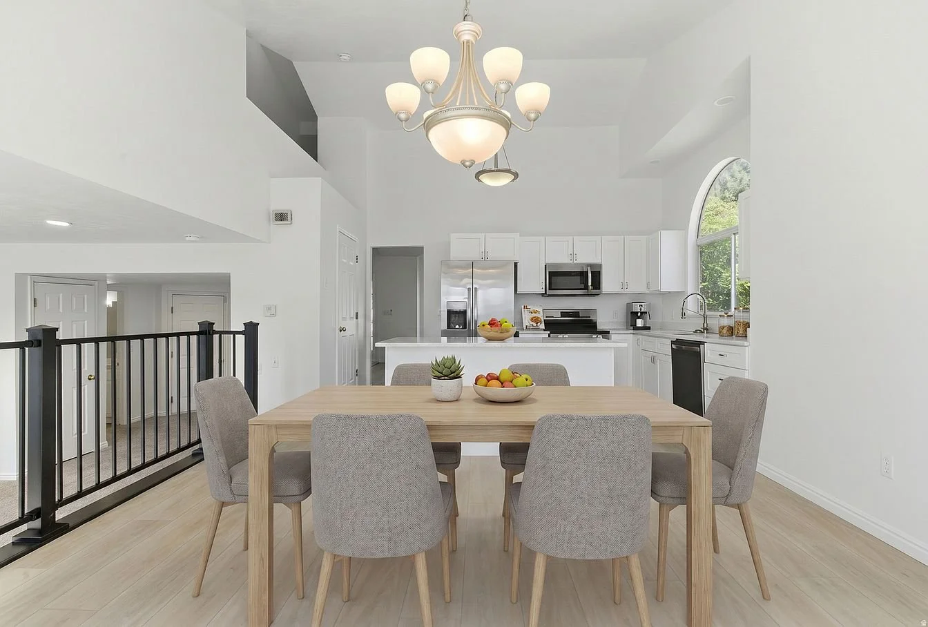 A modern dining area with a wooden table, six gray upholstered chairs, a bowl of fruit, a small potted plant, and a chandelier. Behind, a white kitchen with stainless steel appliances, white cabinets, a window, and a black railing on the left.