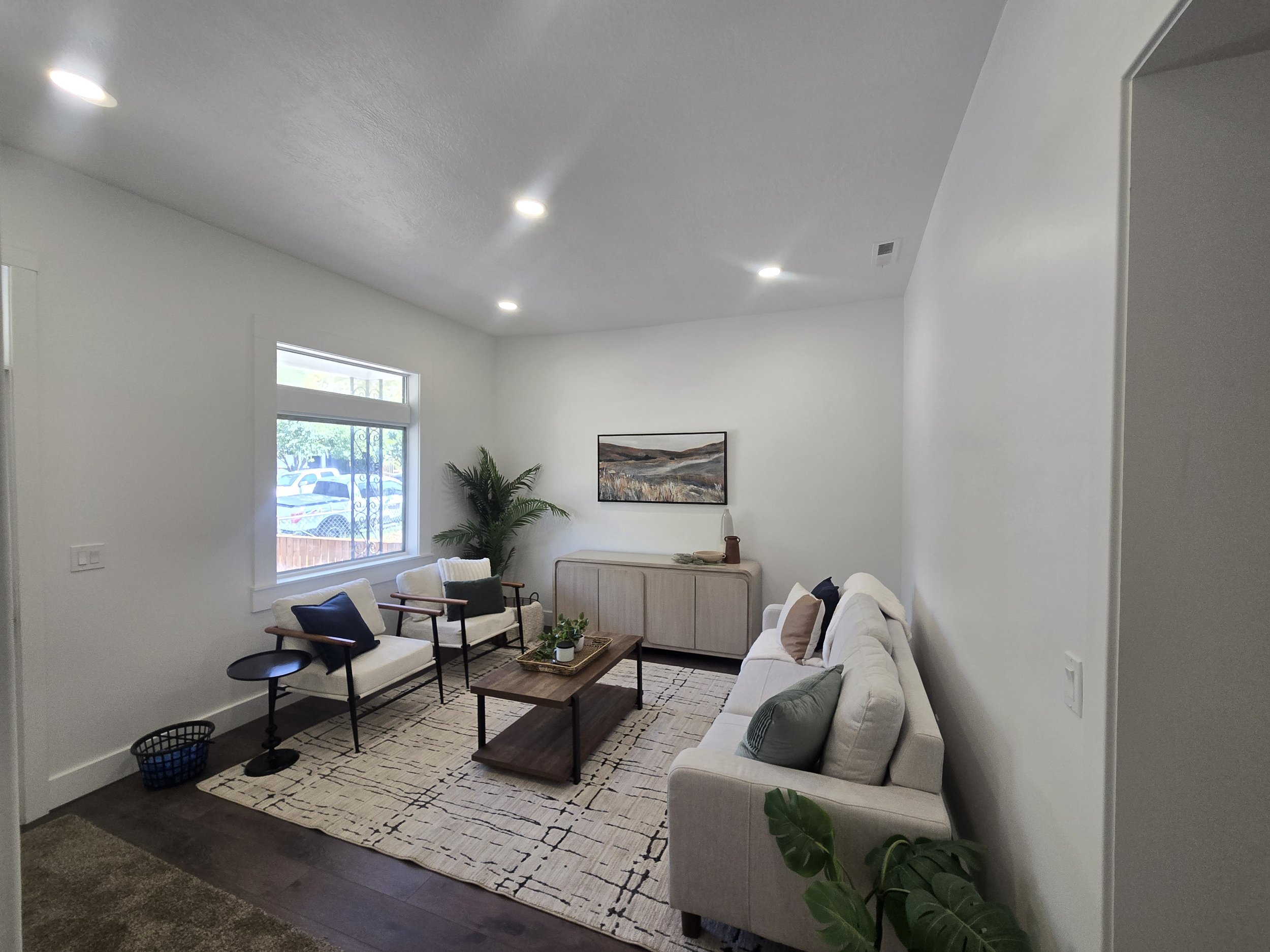 A living room with white walls, a large window, and a ceiling with recessed lighting. The room features a white sofa, black chairs with cushions, a wooden coffee table, a decorative rug, a painting on the wall, a potted plant, and a sideboard with a 