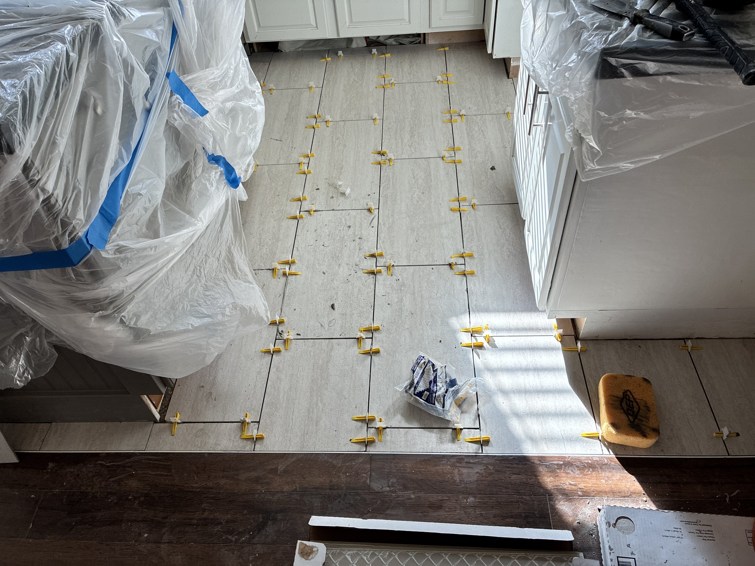Kitchen floor being tiled with yellow spacers and black grout lines. Covered with plastic and construction tools.