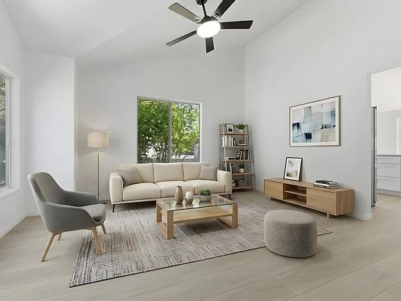 Bright living room with white walls, large window, beige sofa, gray armchair, wooden coffee table, bookshelf, modern artwork, and a ceiling fan.