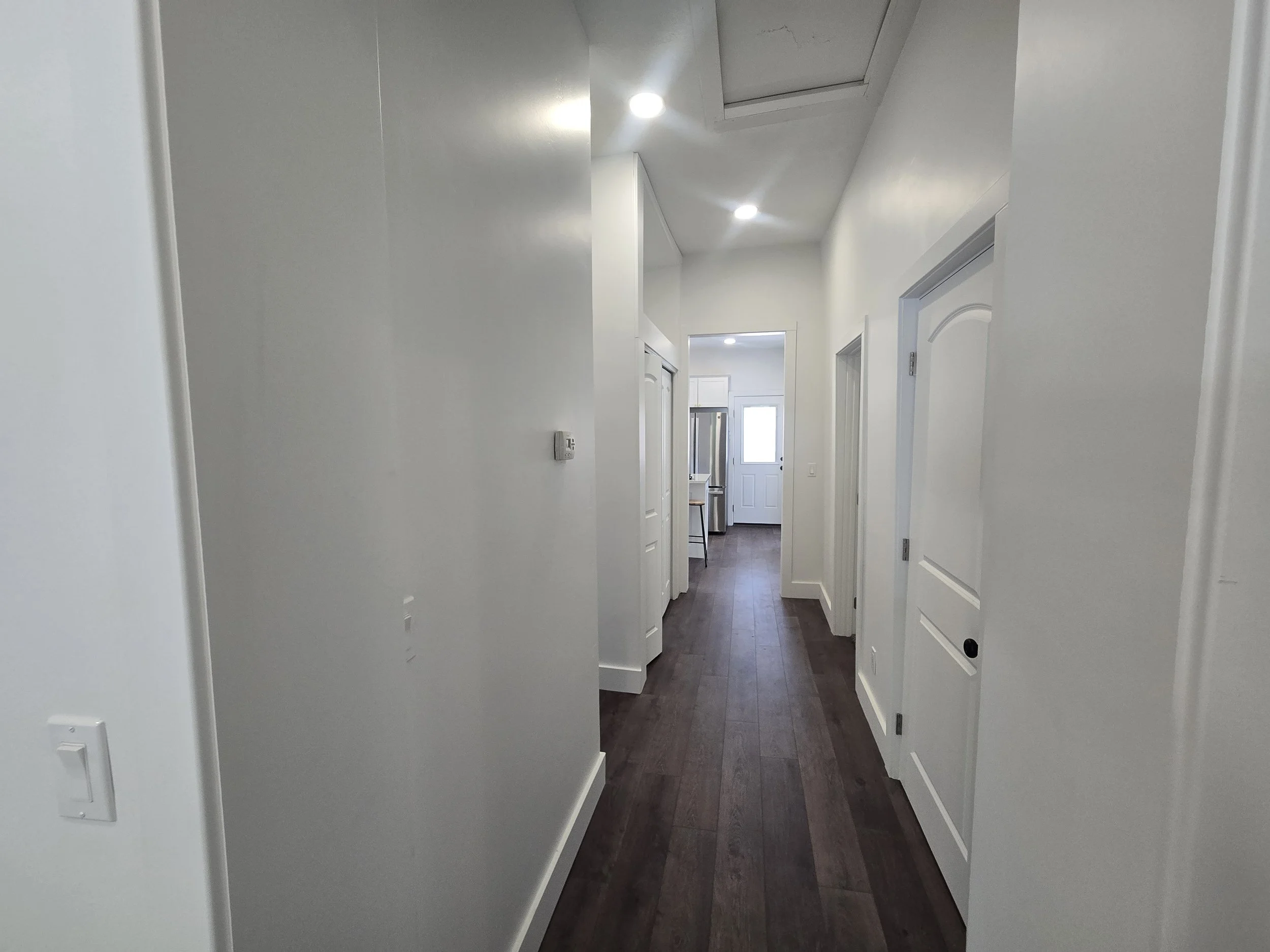 A hallway with white walls, dark hardwood floors, multiple doors, and ceiling lights.