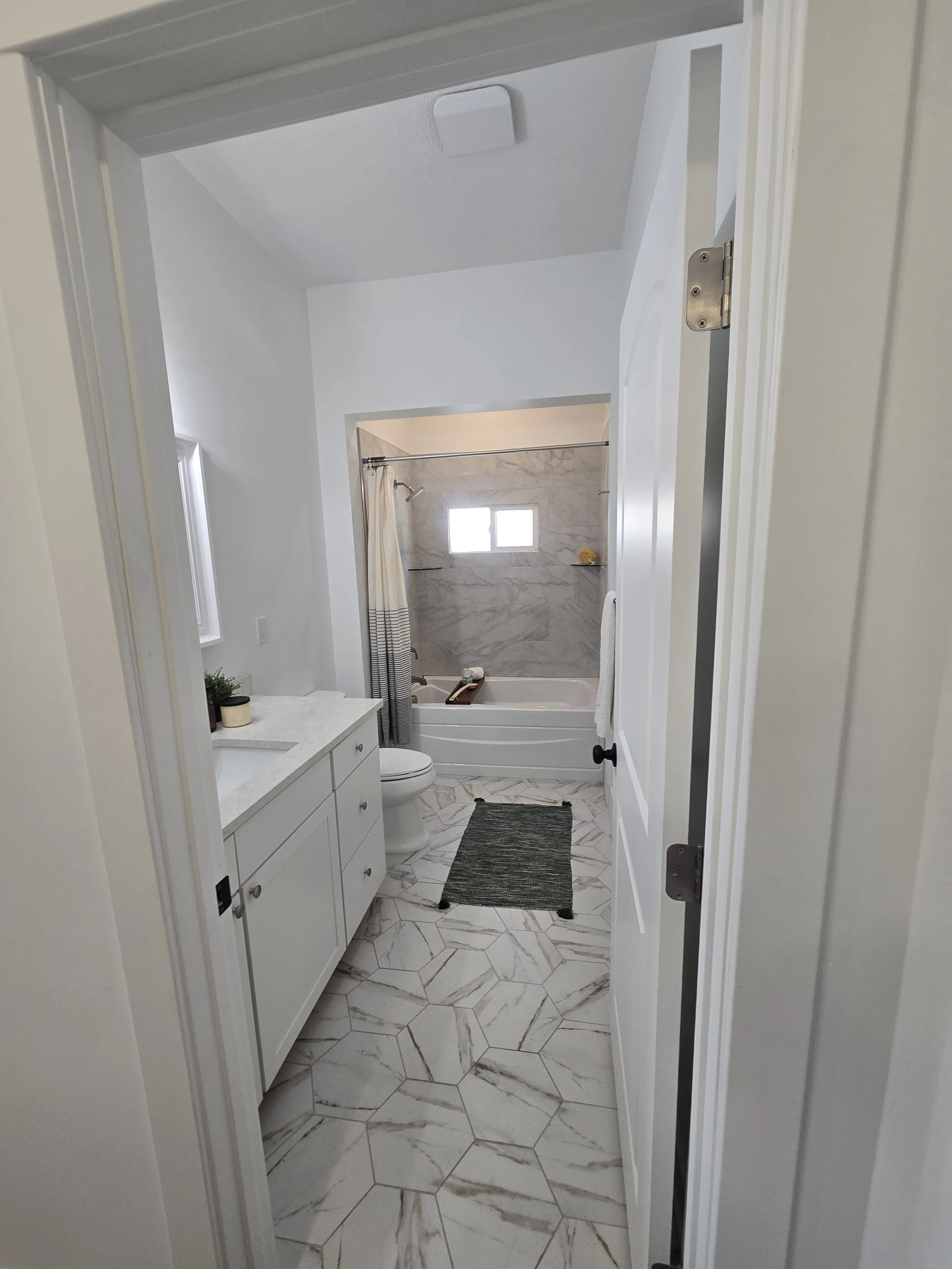 The image shows a bathroom viewed through an open door. The bathroom has white walls, a marble tiled shower area with a small window, a white vanity with drawers, and a toilet. There is a small plant and a candle on the vanity. The floor has large, w