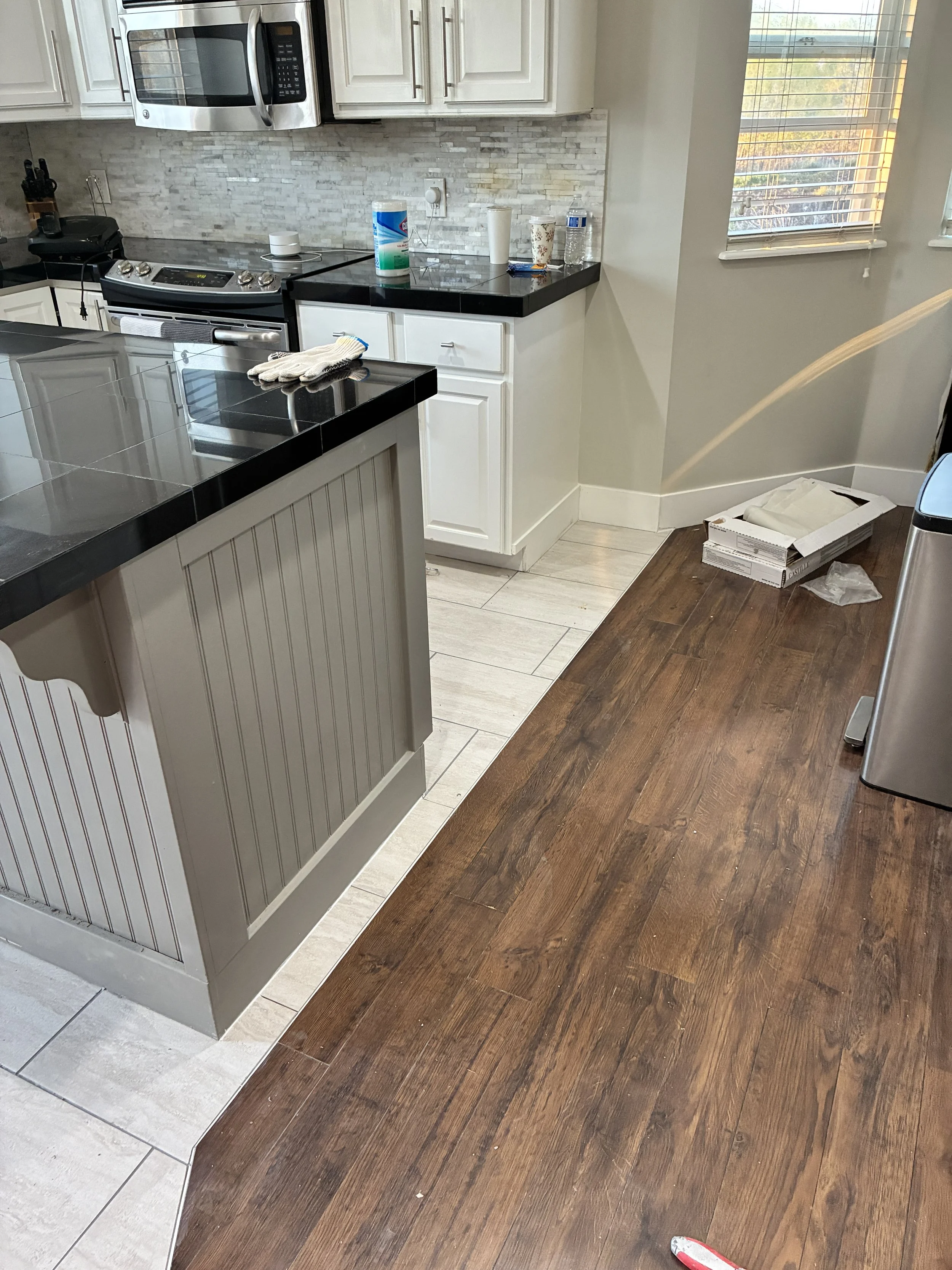 A kitchen with a black countertop, white cabinets, stainless steel microwave, stove, and a gray and white tile backsplash. Items on the counter include a roll of disinfectant wipes, cups, a water bottle, and a snack. On the right, a window with blind