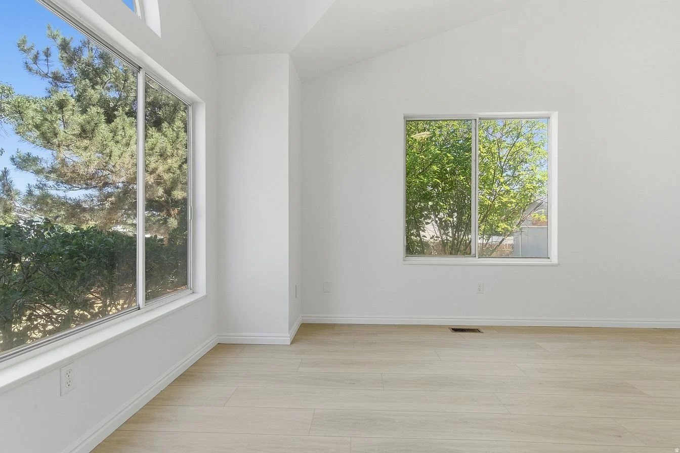 An empty room with light-colored hardwood floors, white walls, and large windows showing trees outside.