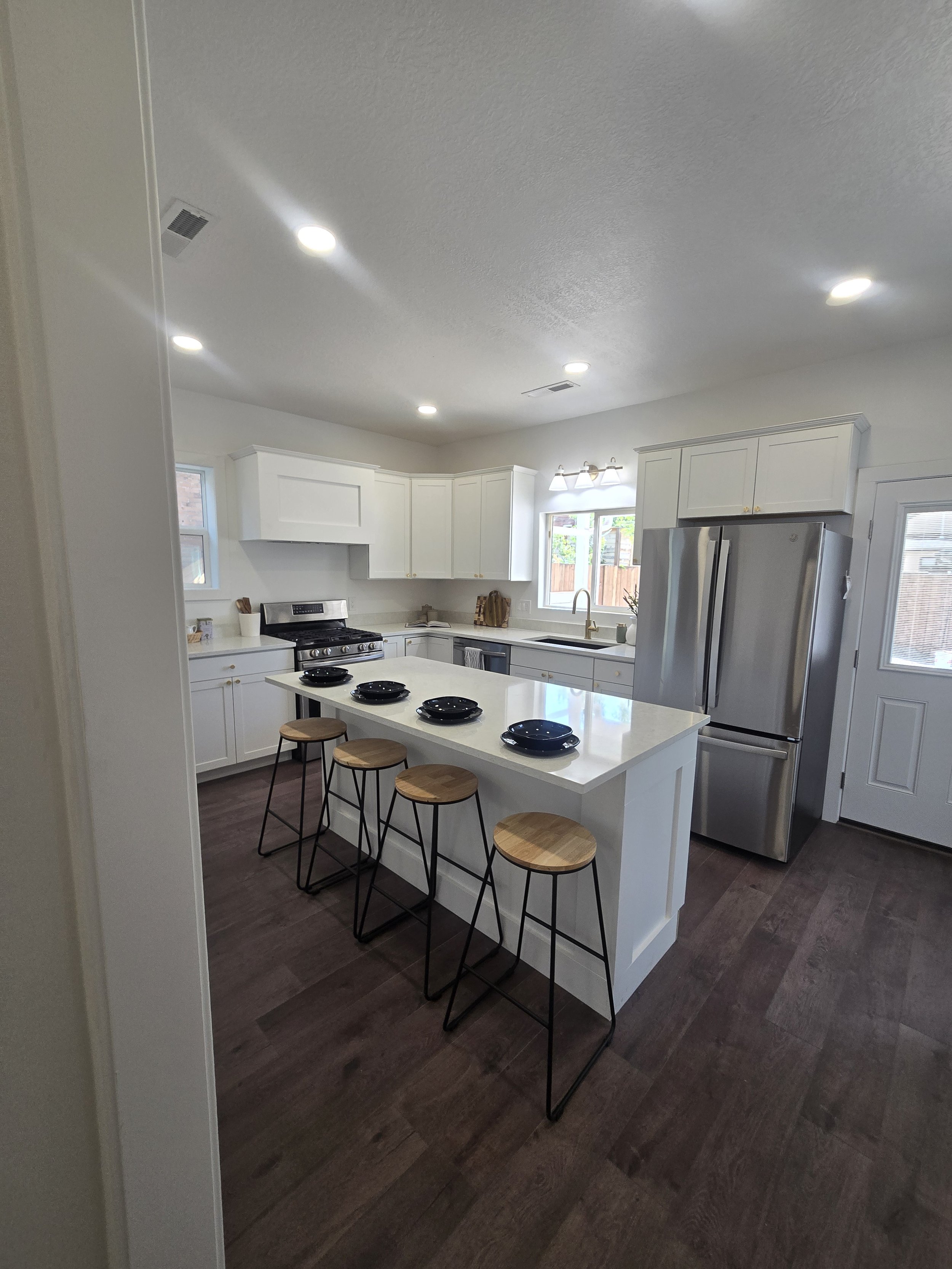 Modern kitchen with white cabinets, stainless steel refrigerator and oven, dark wooden floor, and a white island with black plates and stools.