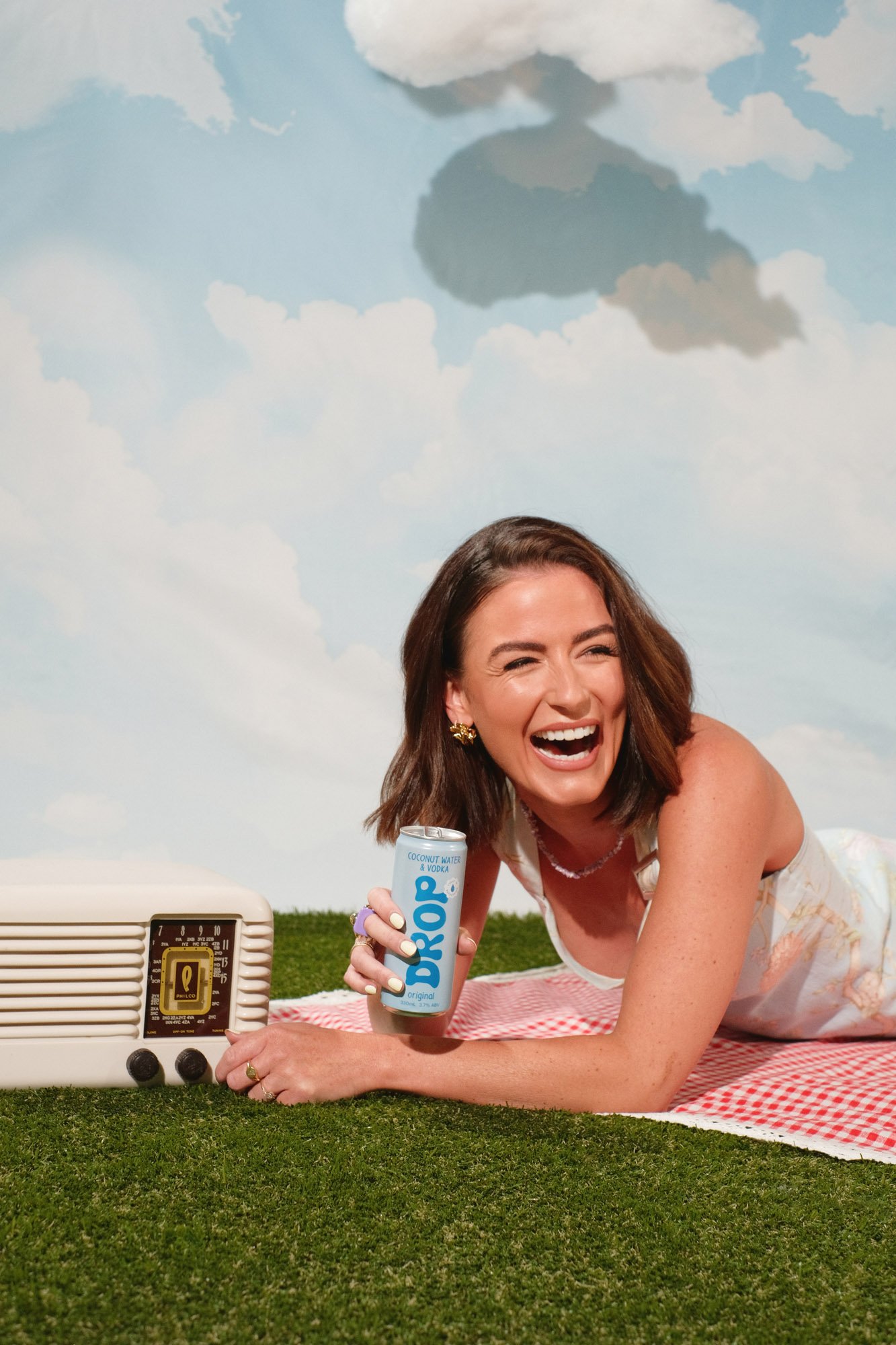 Woman laughing while lying on a red and white checkered blanket outdoors, holding a can of Smirnoff Coconut Water & Vodka, with a vintage portable radio and a cloudy blue sky in the background.