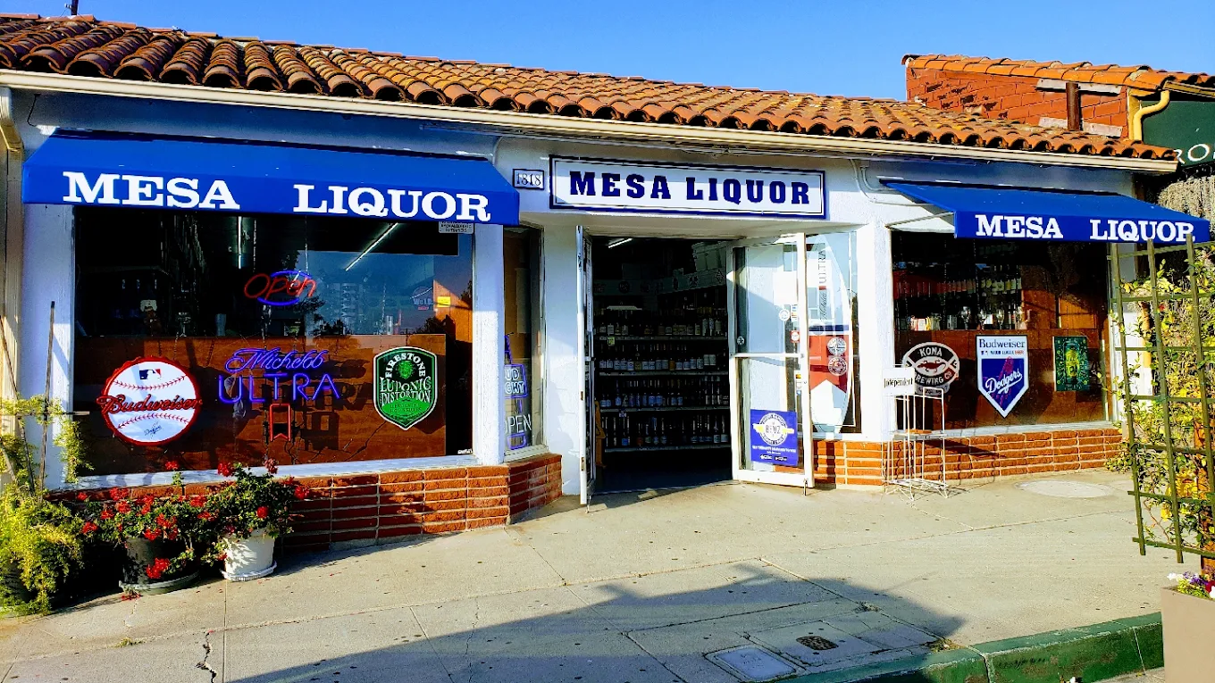 A small liquor store called Mesa Liquor with blue awnings displaying the store name on the storefront. The store has large windows with neon signs for Budweiser, Michelob Ultra, and Green Lizard beer brands. The entrance is open, revealing shelves with bottles inside. There are flower pots and plants outside, and the building has a tiled roof with a blue sky overhead.