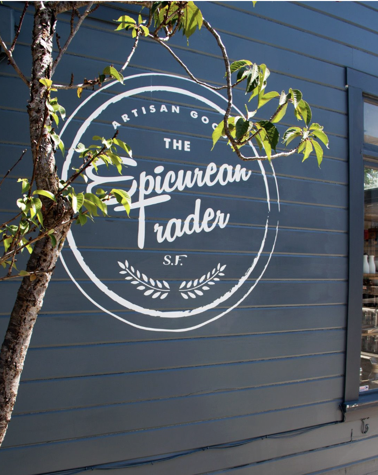 Sign on a building says 'The Epicurean Trader' with the slogan 'Artisan Goods' and 'S.F.' underneath, surrounded by green leaves and a tree branch.