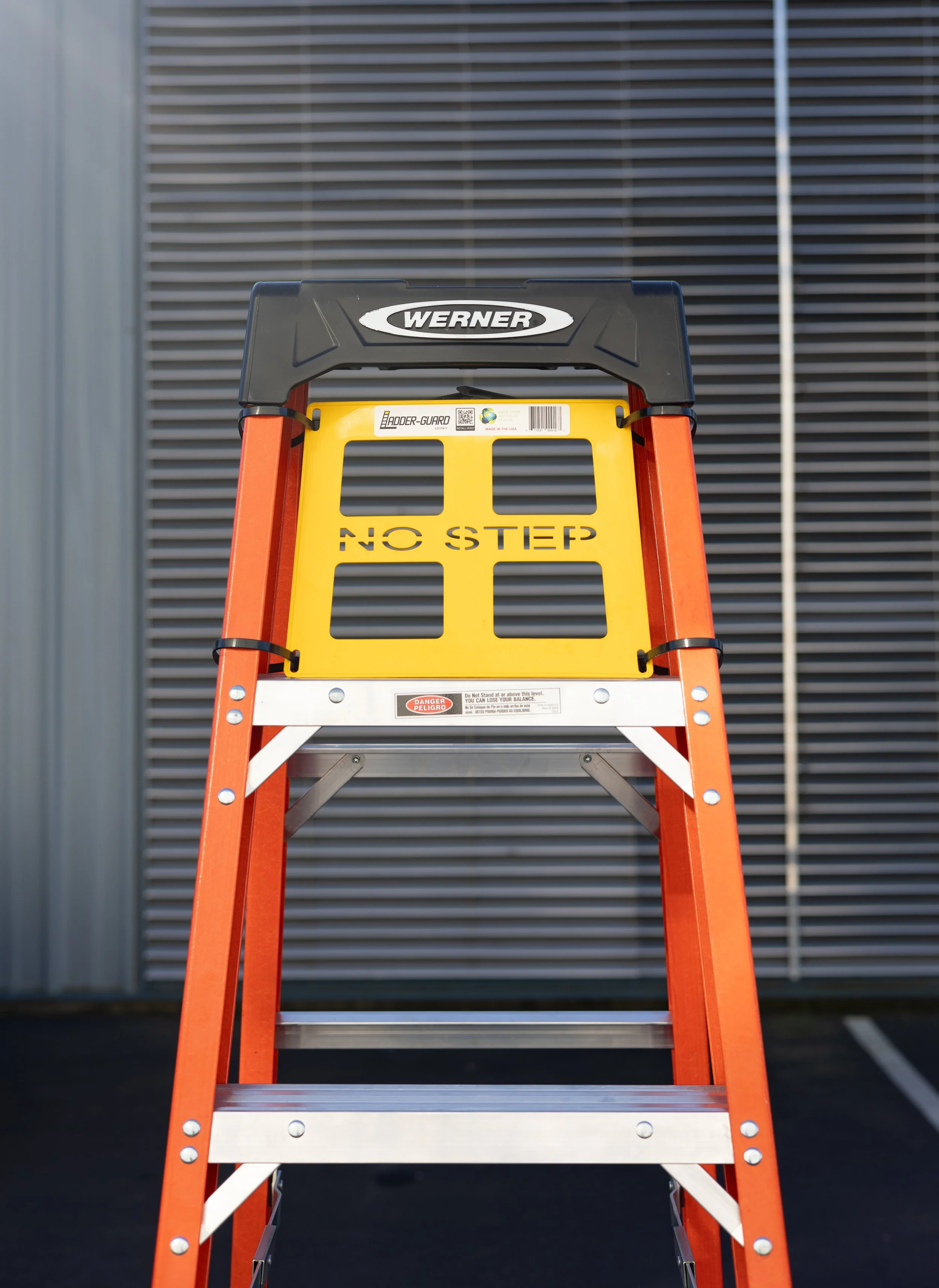 A orange and silver stepladder with a black top and yellow backer guard, positioned in front of a dark gray wall with metallic slats.