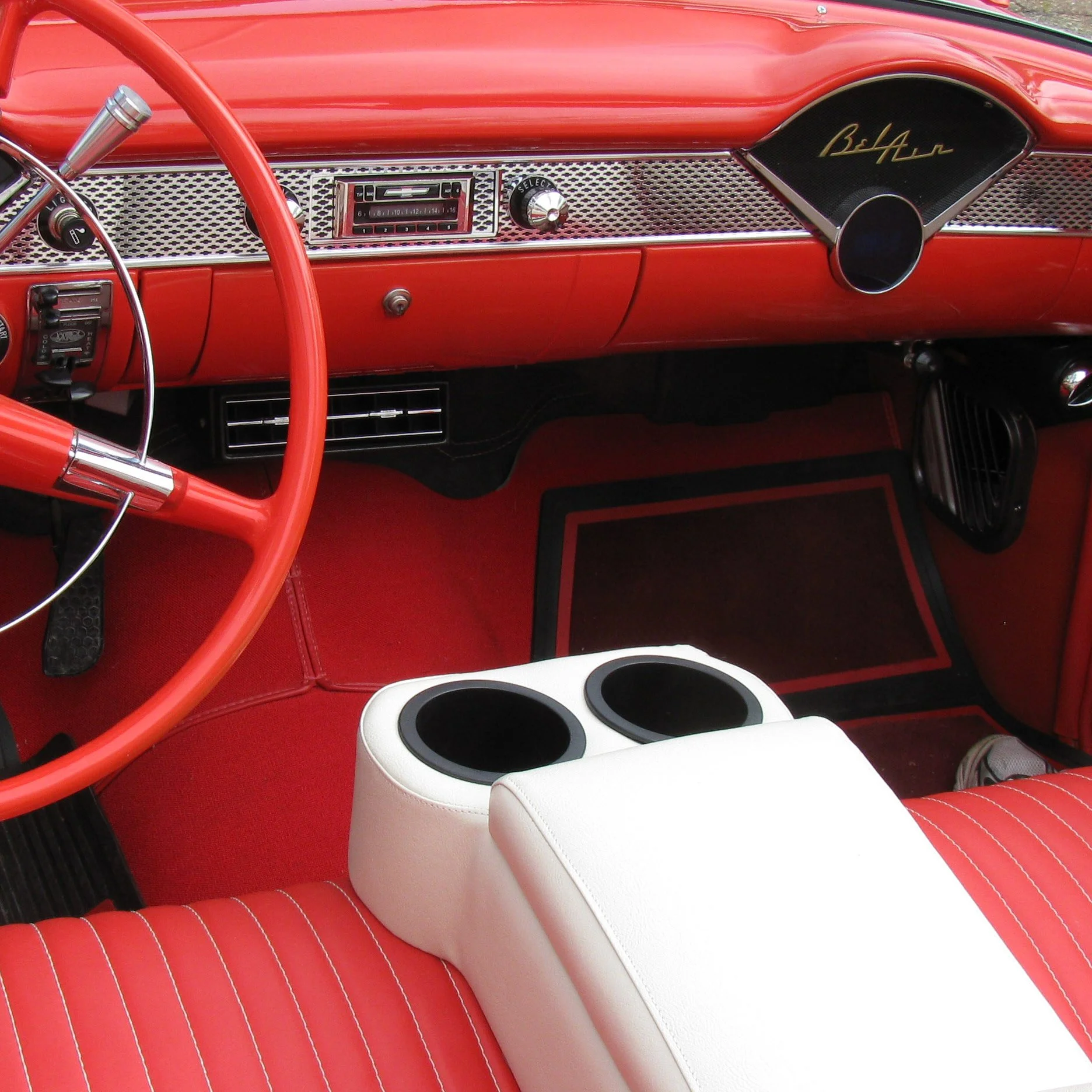 Interior of a vintage red car with a red steering wheel, white and red seating, black foot pedals, and a white center console with black cup holders.