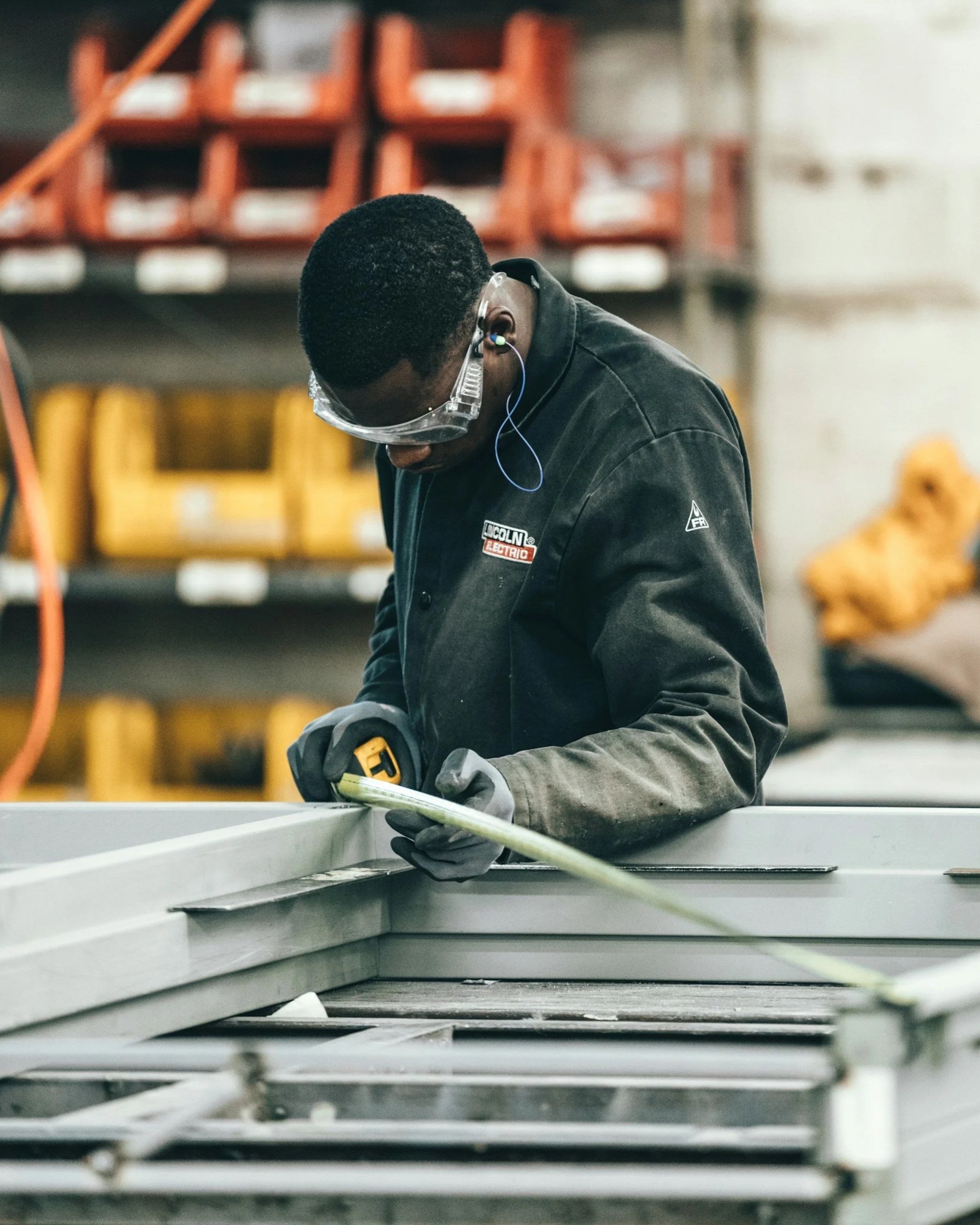 A worker with safety glasses and earplugs, wearing a black jacket, is working on a metal surface in an industrial warehouse with yellow chairs and orange storage bins in the background.
