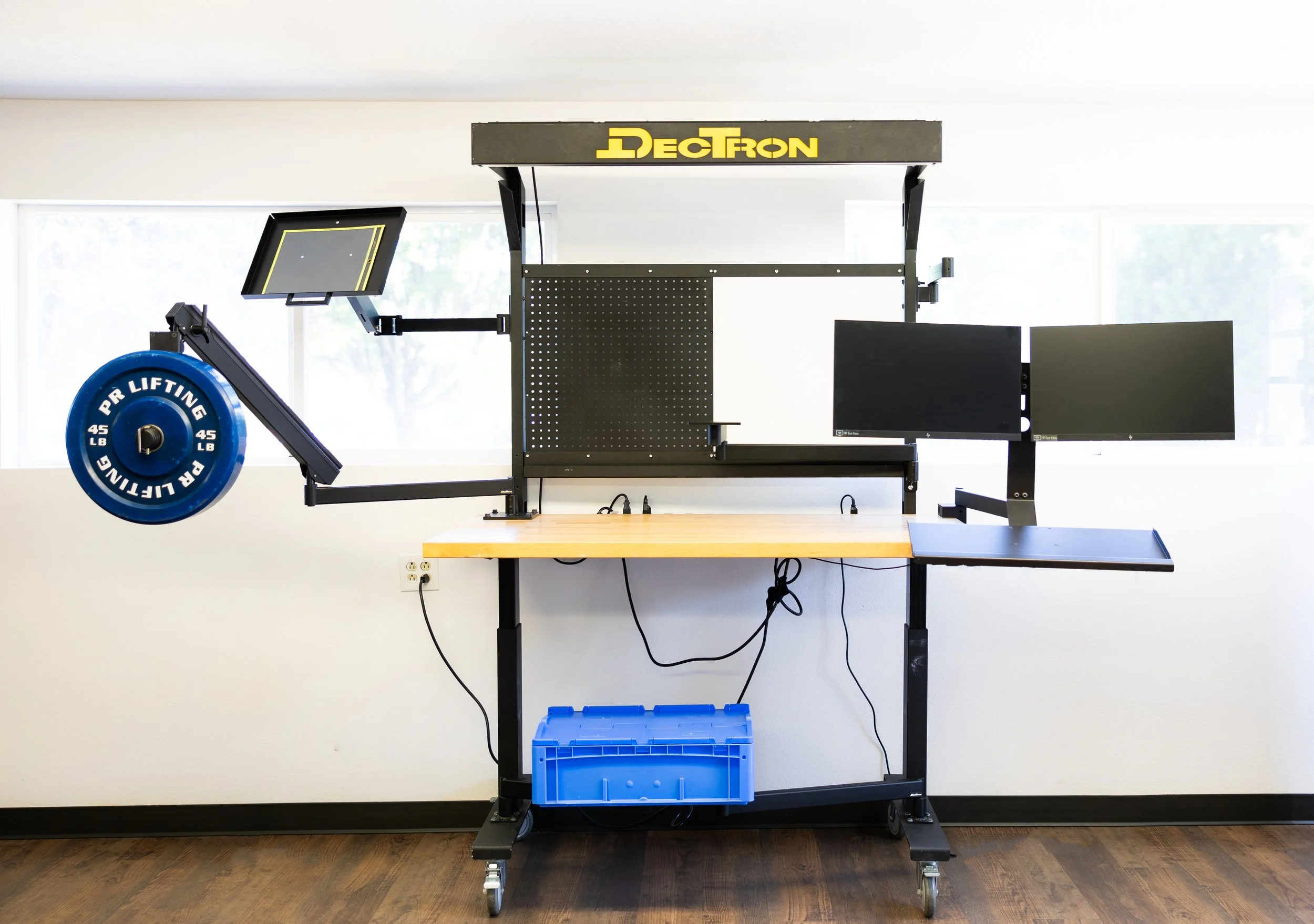 A mechanical workstation with a black pegboard, monitors, and a blue weight plate, labeled 'PR Lifting 45 LB'.