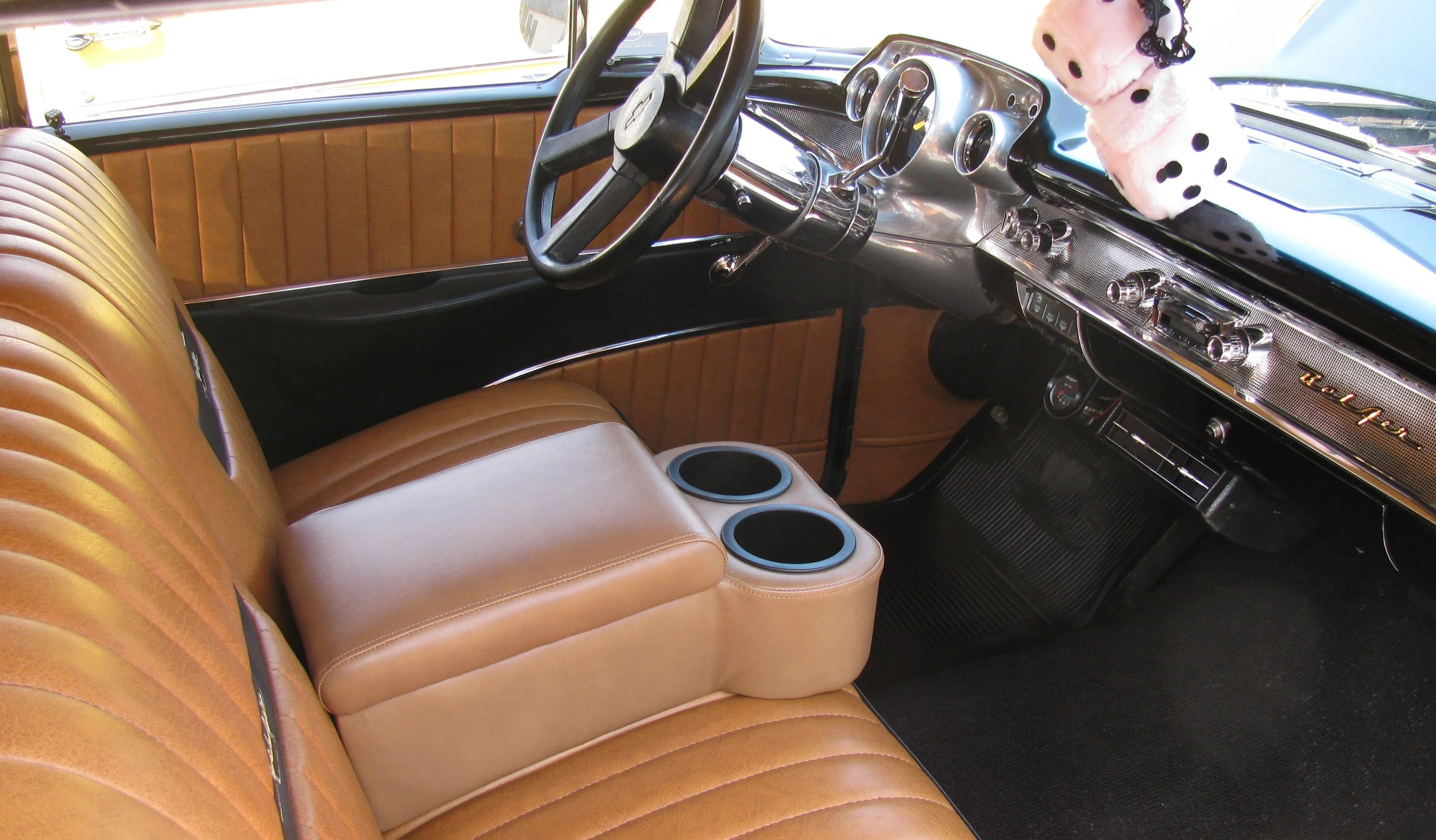 Interior of a classic car showing tan leather bench seats, a steering wheel, and a dashboard with various controls, with fuzzy pink dice hanging from the rearview mirror.