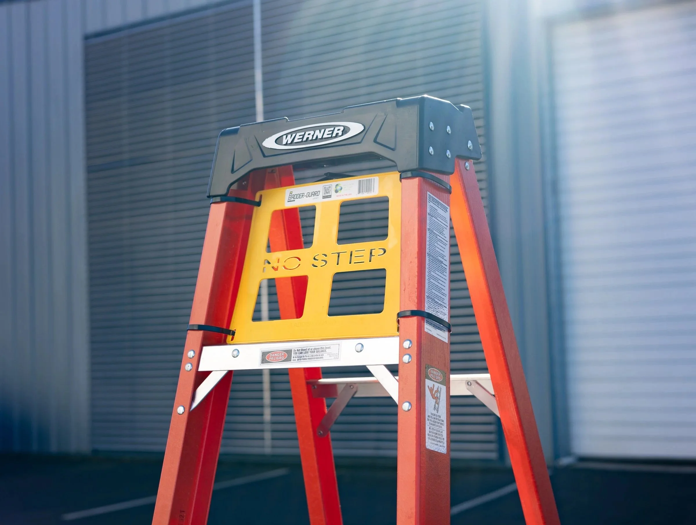 Orange and yellow step ladder with a black safety top, in front of a building with metal siding, with warning labels on the ladder.