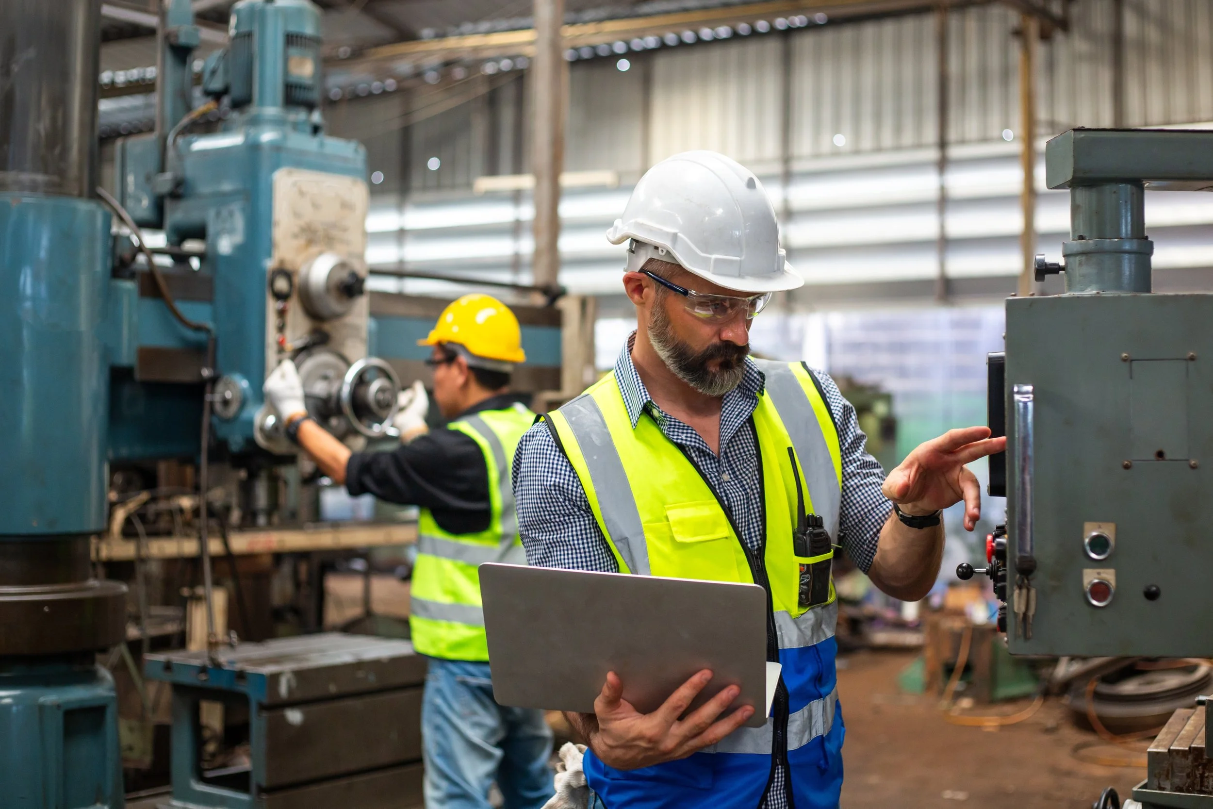 Two industrial workers wearing safety helmets and high-visibility vests working with machinery in a factory.