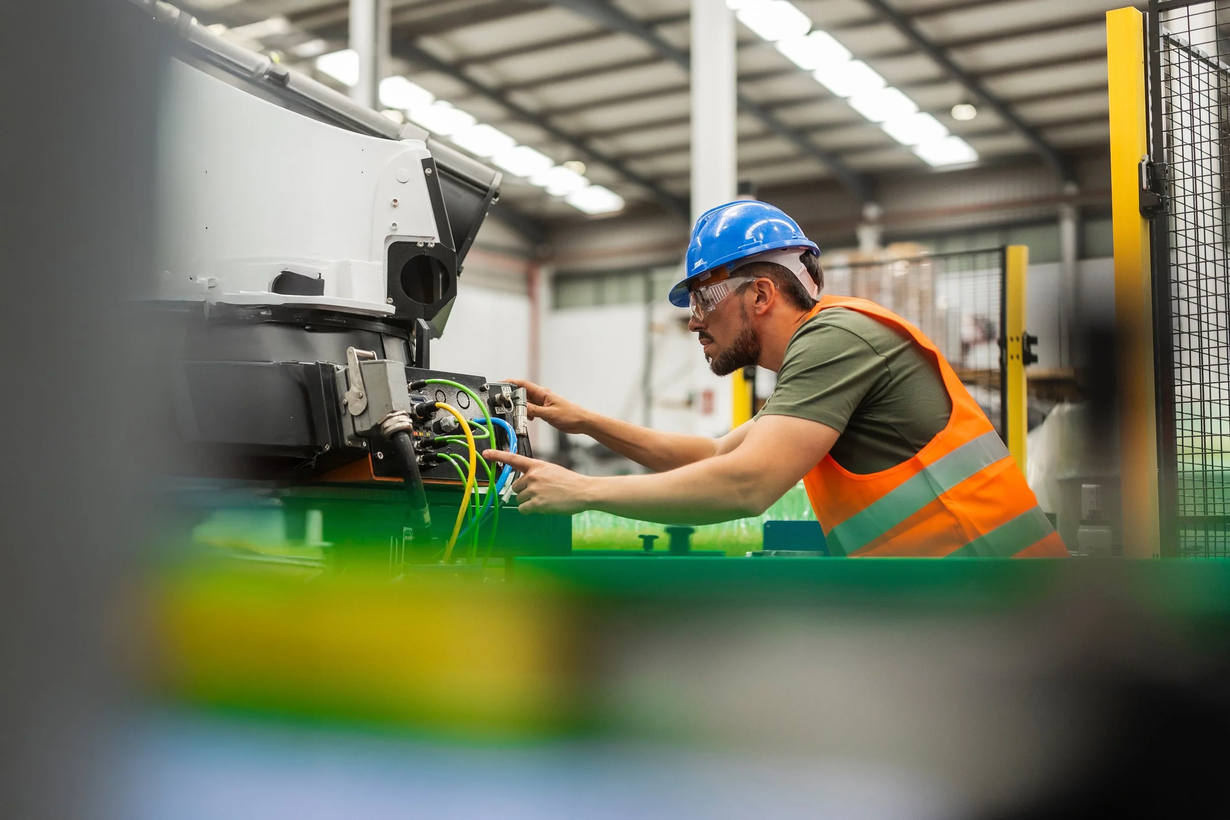 A technician wearing a blue safety helmet, goggles, and an orange safety vest working on electrical equipment in an industrial setting with a warehouse roof in the background.