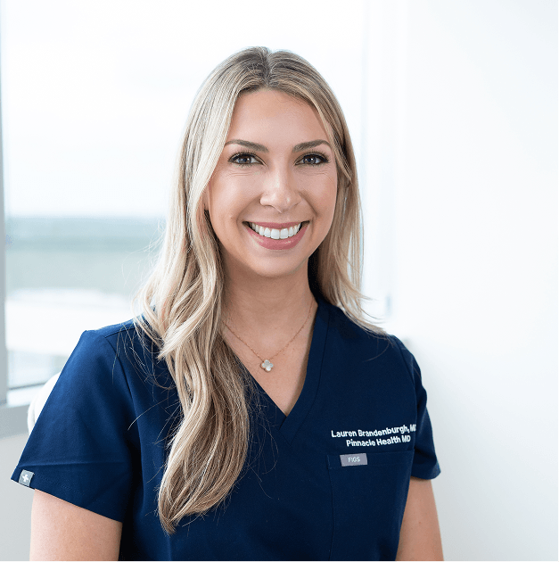 A smiling woman with long blonde hair wearing navy medical scrubs with the name tag 'Lauren Brandenburg, NP Pinnacle Health MD' in a bright, clinical setting.