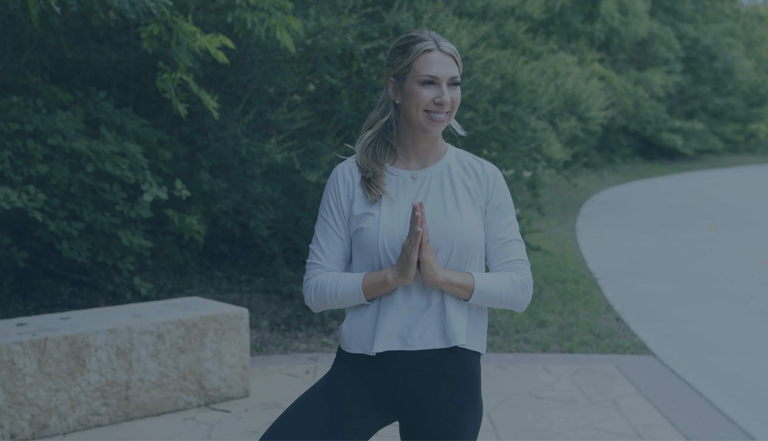 A woman in a white top and black pants practicing yoga outdoors with her hands in prayer position, smiling amidst green trees and a curved pathway.