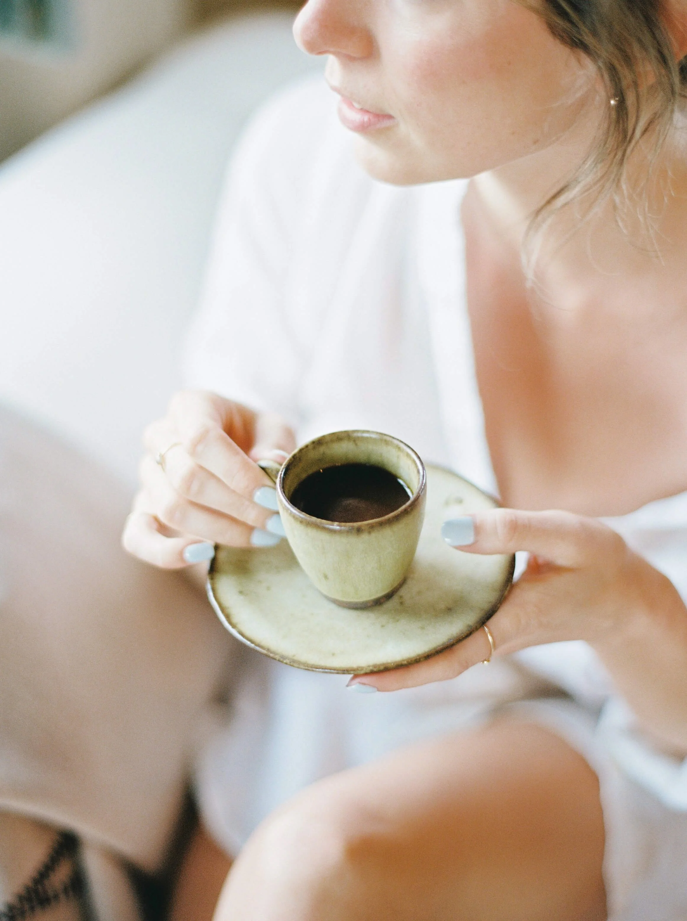 A person holding a tea cup with dark beverage on a small matching saucer, wearing a white shirt and a gold ring on their finger, with painted nails.