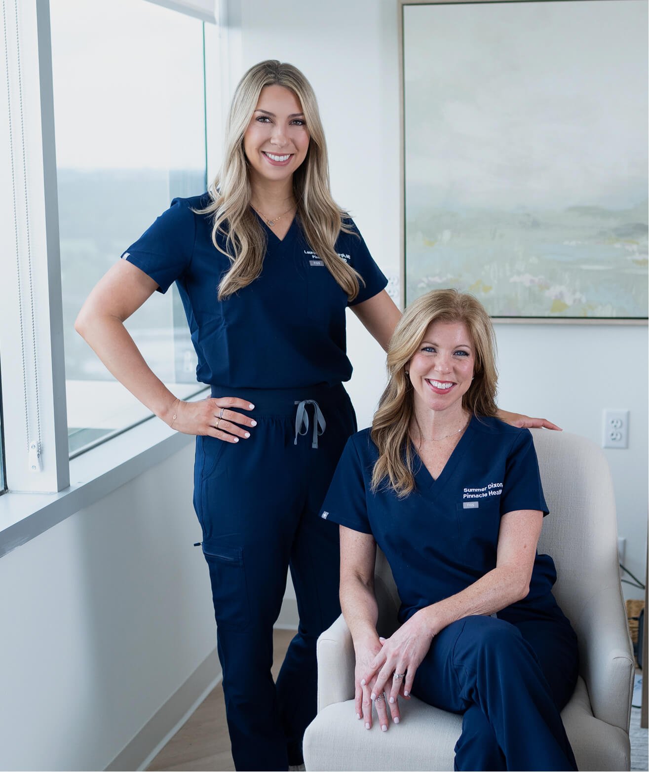 Two women in navy scrubs, one standing and one sitting indoors at a medical facility. The standing woman has blonde hair, and the seated woman has reddish hair. Both are smiling and appear to be healthcare professionals.