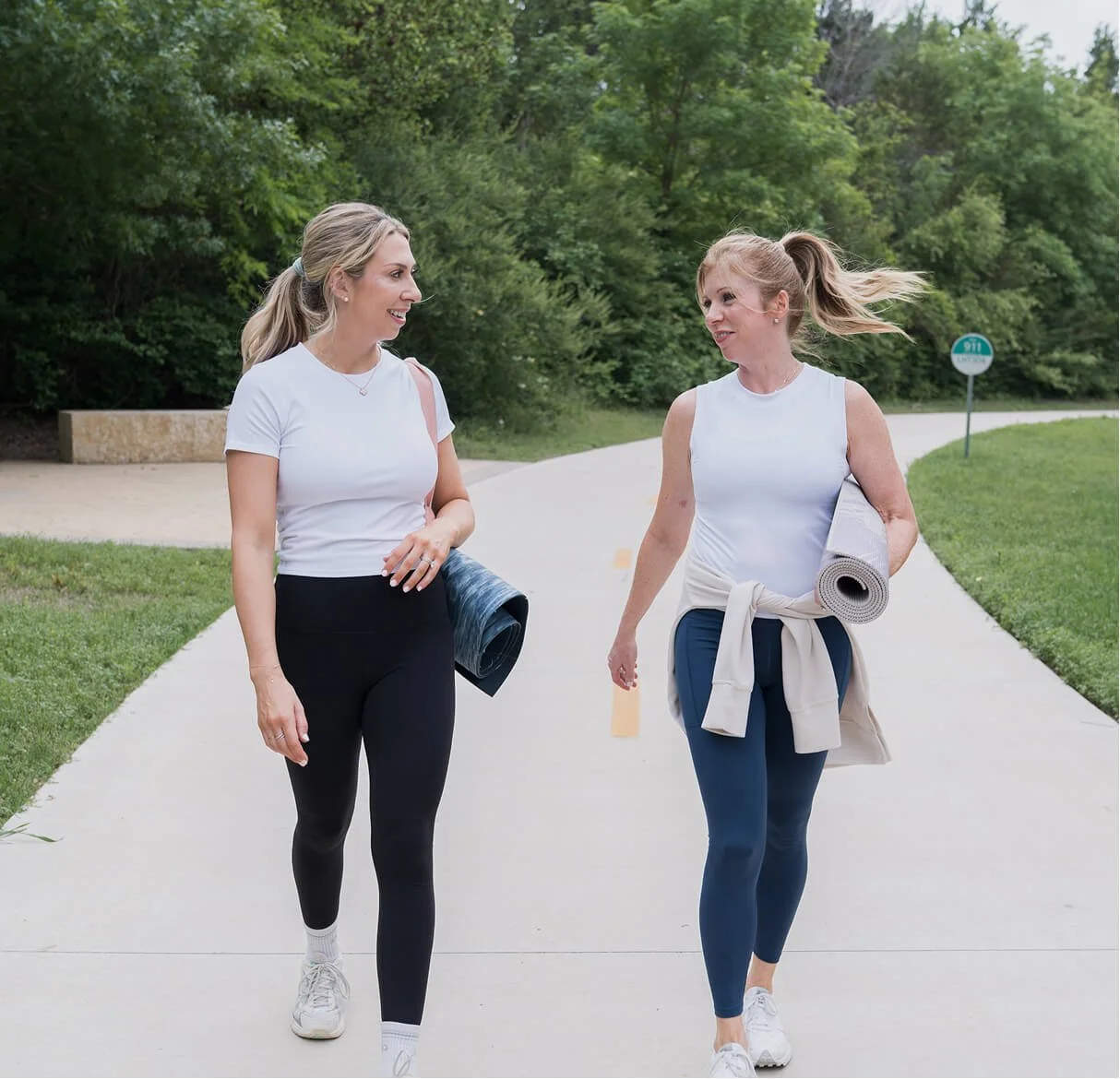 Two women walking outdoors on a paved path, carrying yoga mats, dressed in athletic wear, and smiling while engaging in conversation, with greenery and trees in the background.