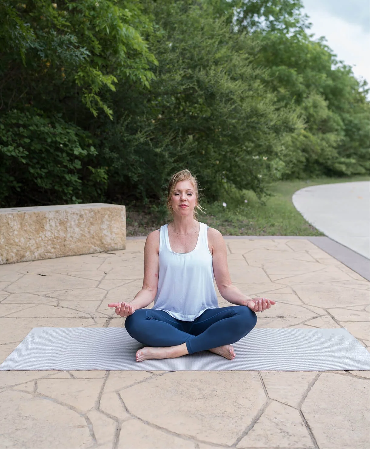 A woman meditating outdoors on a yoga mat on a stone patio surrounded by green trees, sitting cross-legged with hands resting on her knees in a mudra position.