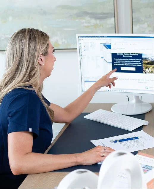A woman in medical scrubs sitting at a desk, pointing at a computer screen with a calendar or scheduling software displayed.