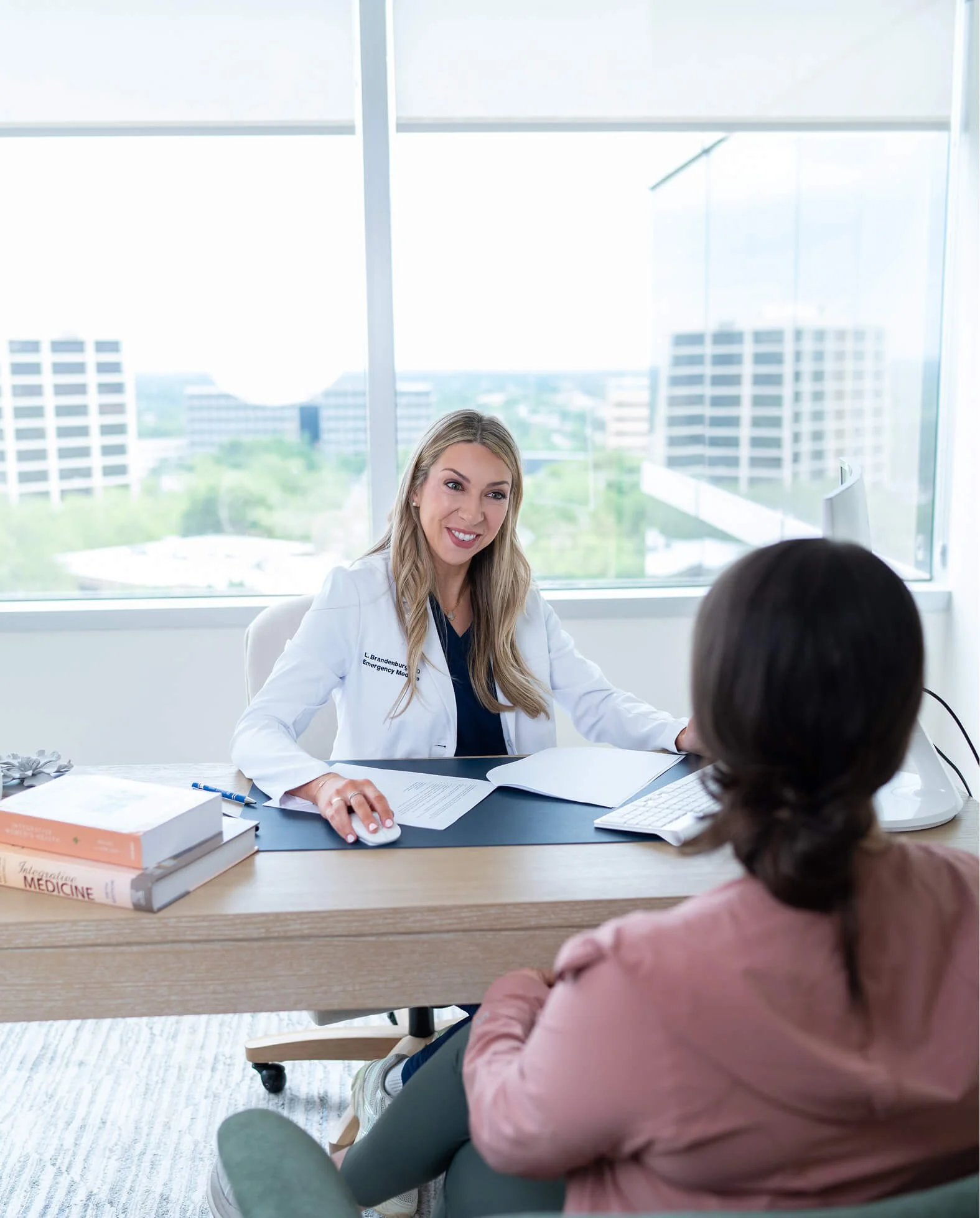 A female doctor or healthcare professional with long blonde hair, wearing a white coat, is smiling and talking to a patient during an appointment in a modern hospital or clinic. The patient, a woman with dark hair, sits across the desk. The office has large windows with city views.