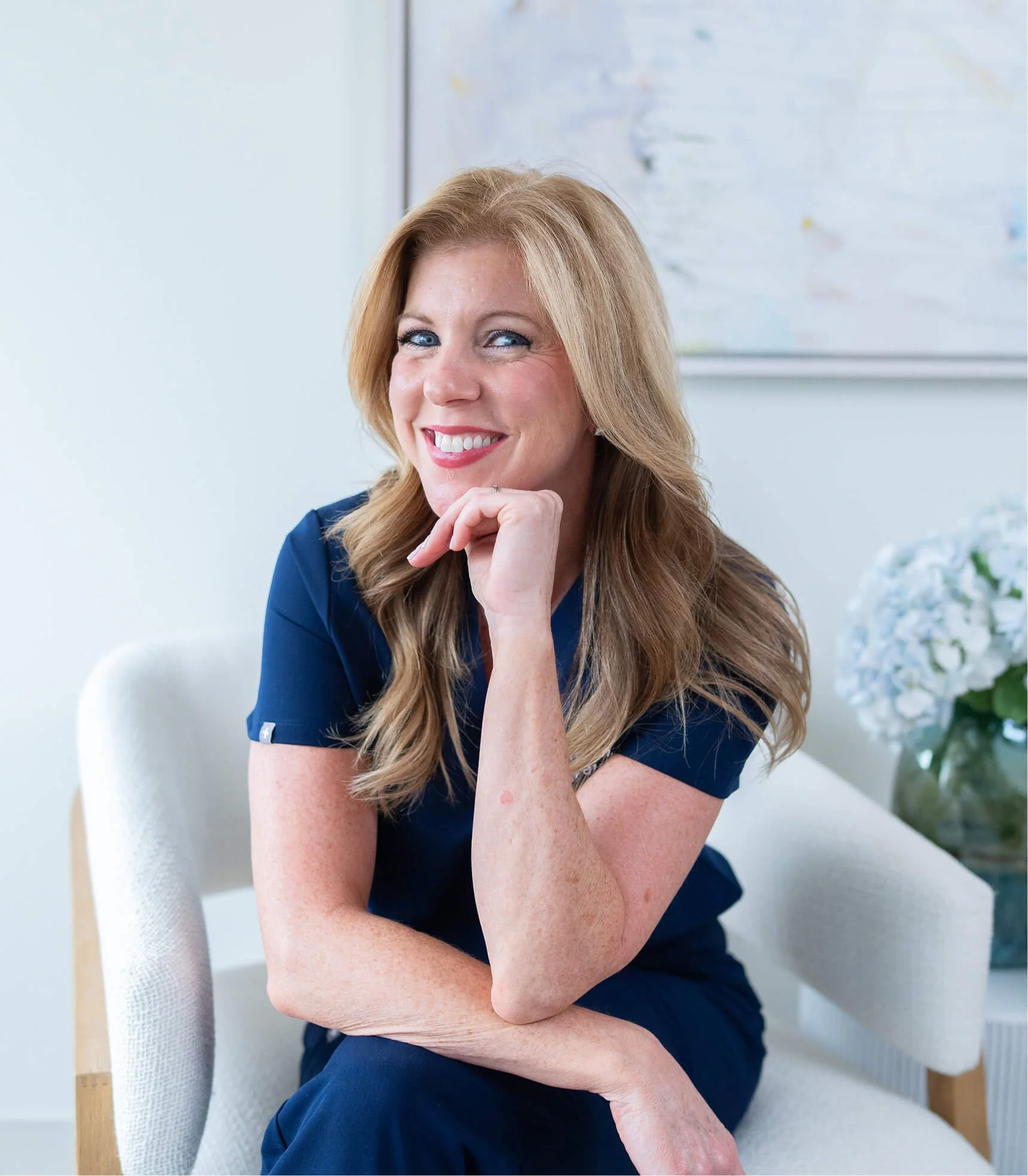 A woman with long red hair and blue eyes smiling while sitting on a white chair in a bright room. She is wearing a navy blue top and resting her chin on her hand.