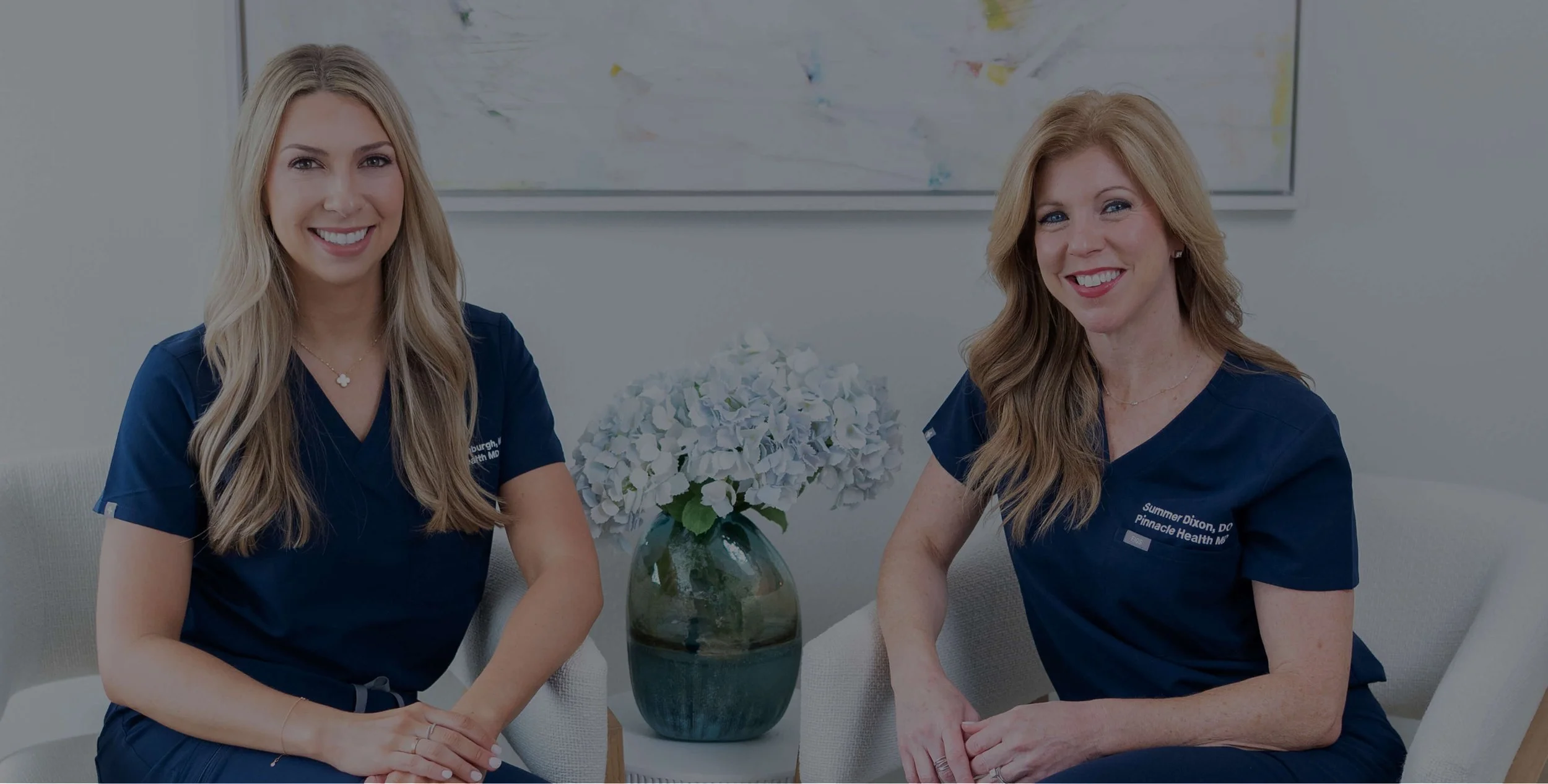 Two women wearing navy medical scrubs sitting at a table with a vase of white hydrangeas, smiling at the camera.