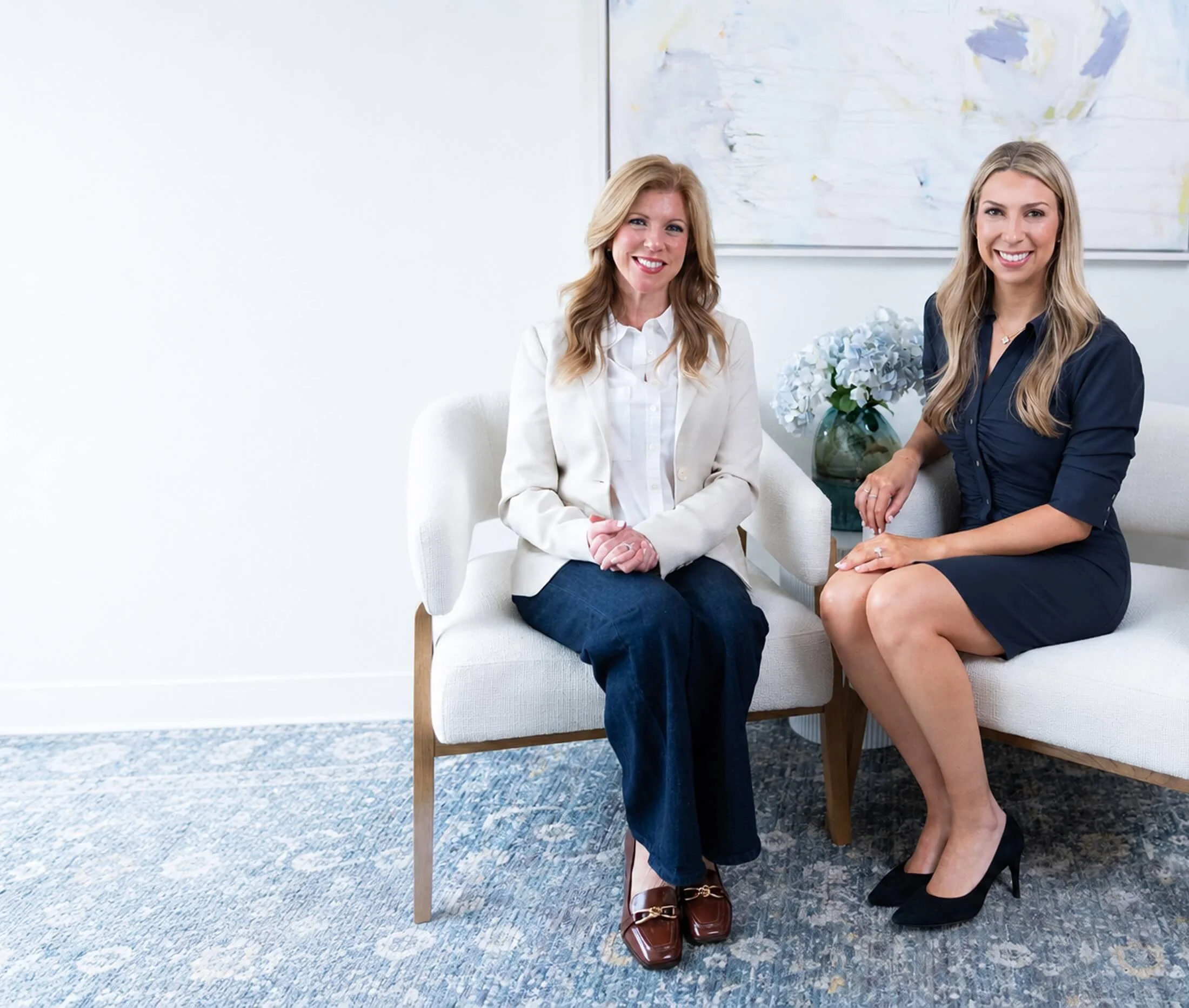 Two women sitting on a white couch in a room with a large abstract painting on the wall, a bowl of white flowers, and a blue and white patterned carpet.