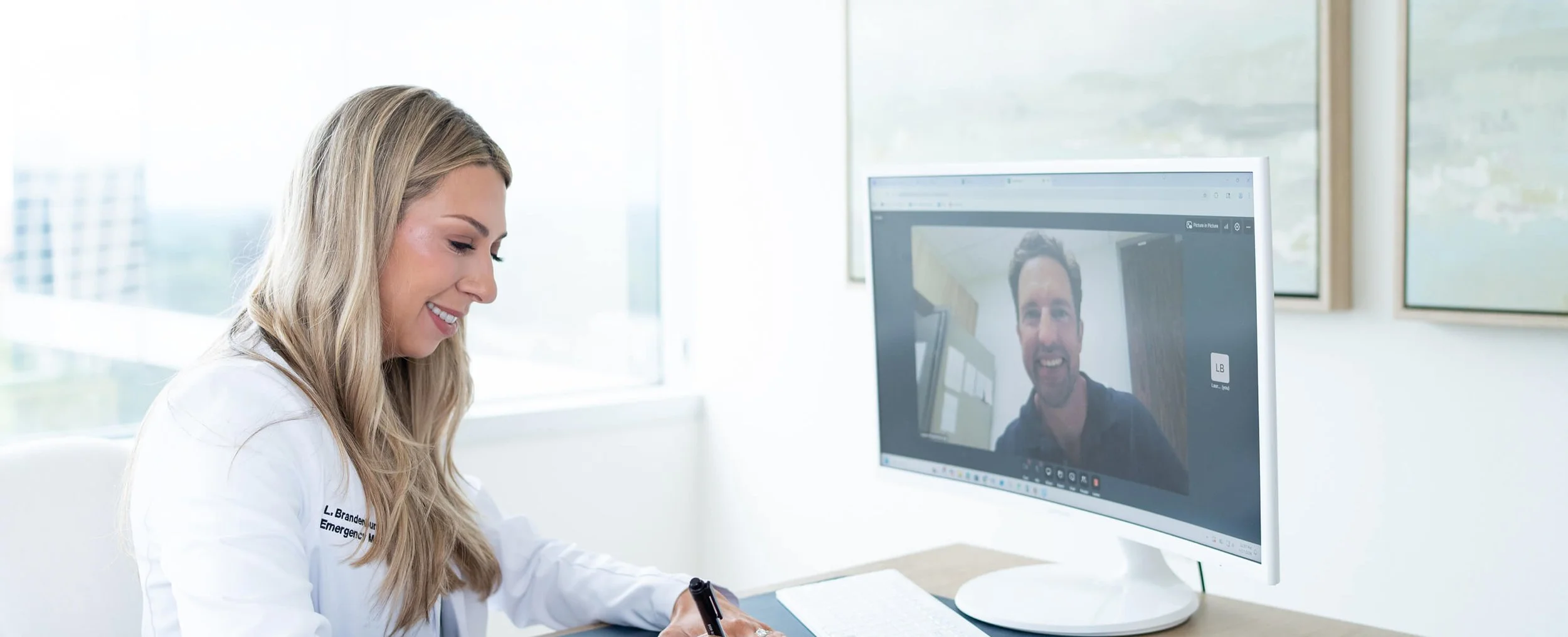 A female healthcare professional in a white coat smiling during a video call with a male colleague on a computer monitor in a bright, modern office.