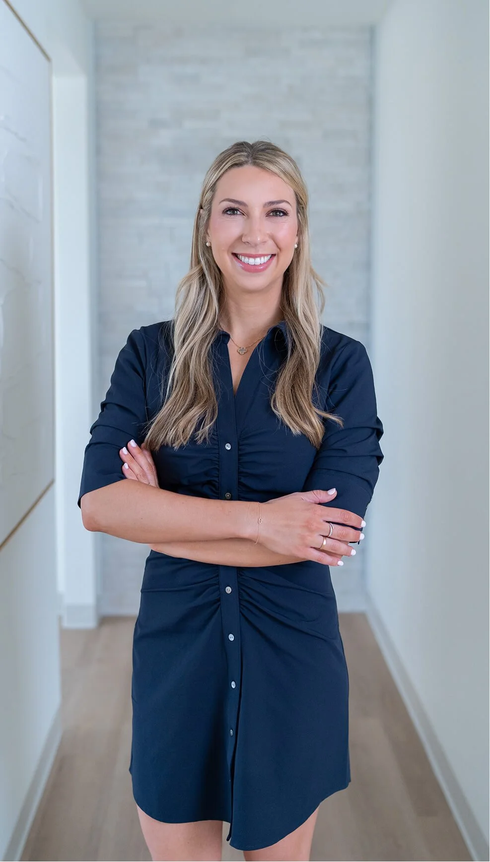 A smiling woman with blonde hair, wearing a dark blue dress, standing with arms crossed in a modern, light-colored hallway.