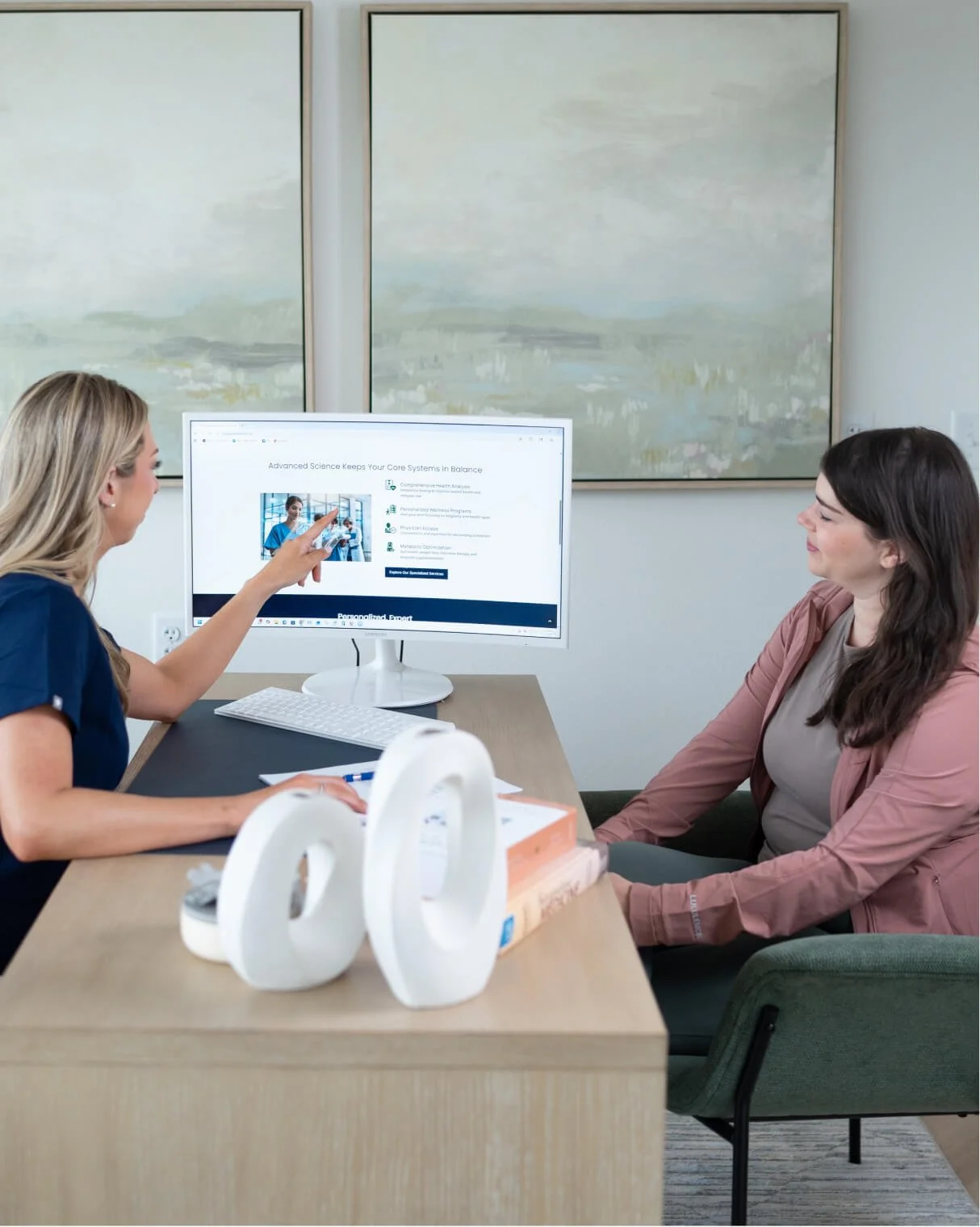 Two women having a meeting in an office, one explaining something on a computer screen to the other. The office has abstract paintings on the wall, a desk with books, a white decorative sculpture, and a computer with a large monitor.